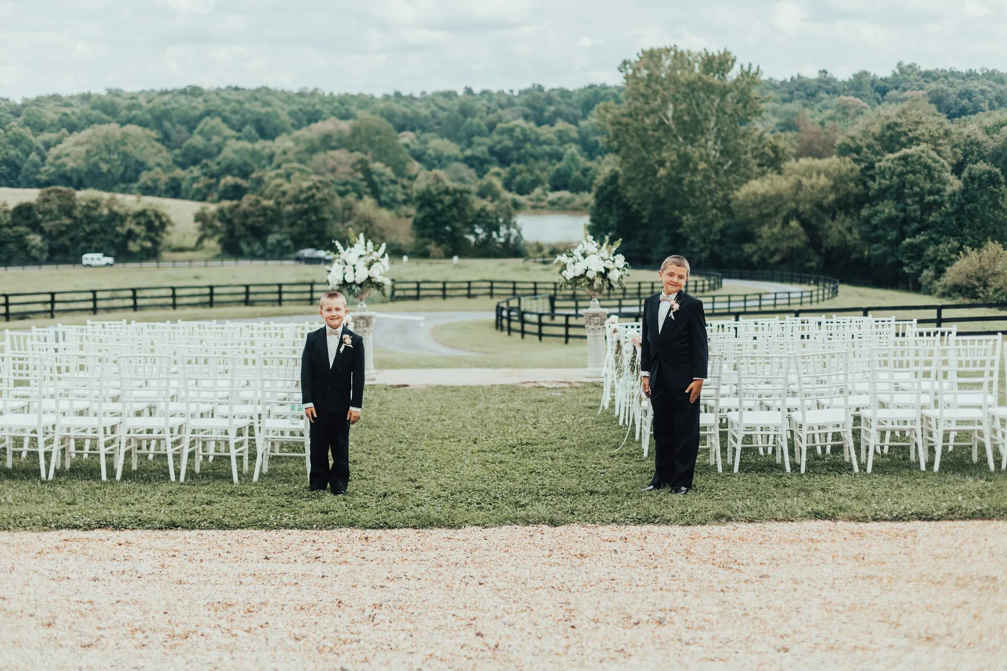 Two grooms in black suits stand at wedding altar with white chairs and floral arch overlooking rural Virginia countryside.
