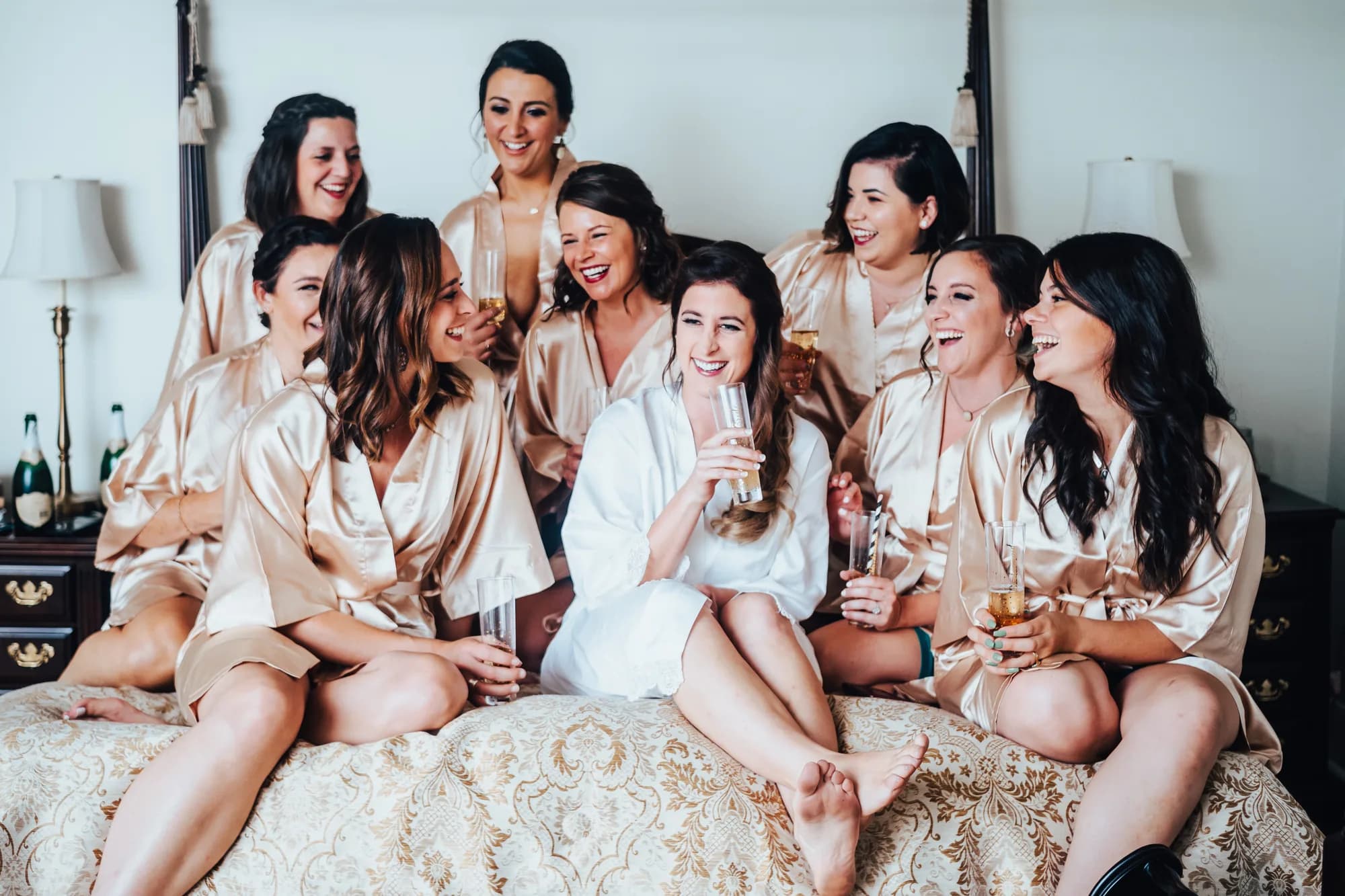 Bride and bridesmaids in satin robes laughing together on a bed with champagne before the wedding