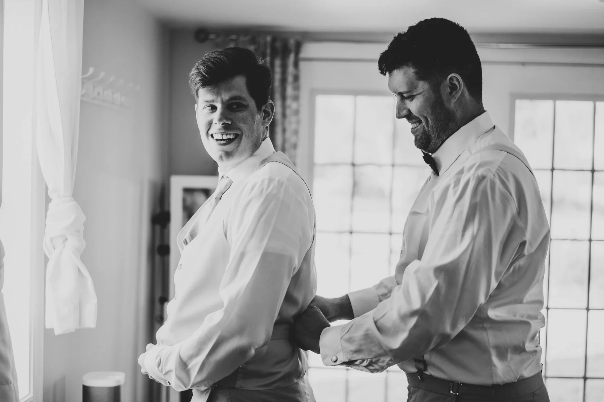 Groom laughing as groomsman helps fasten his cufflinks while getting ready before the wedding