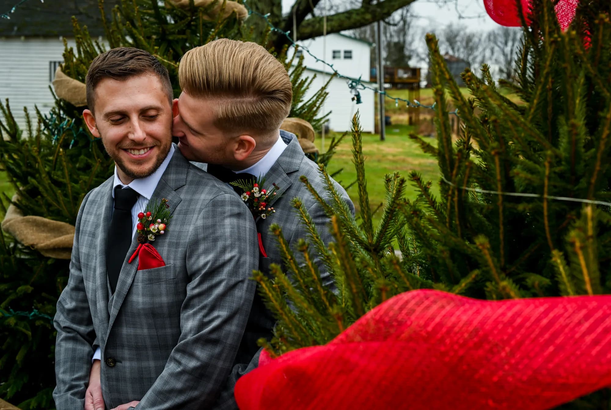 Two grooms in matching plaid suits share a tender kiss among Christmas trees at an outdoor winter wedding