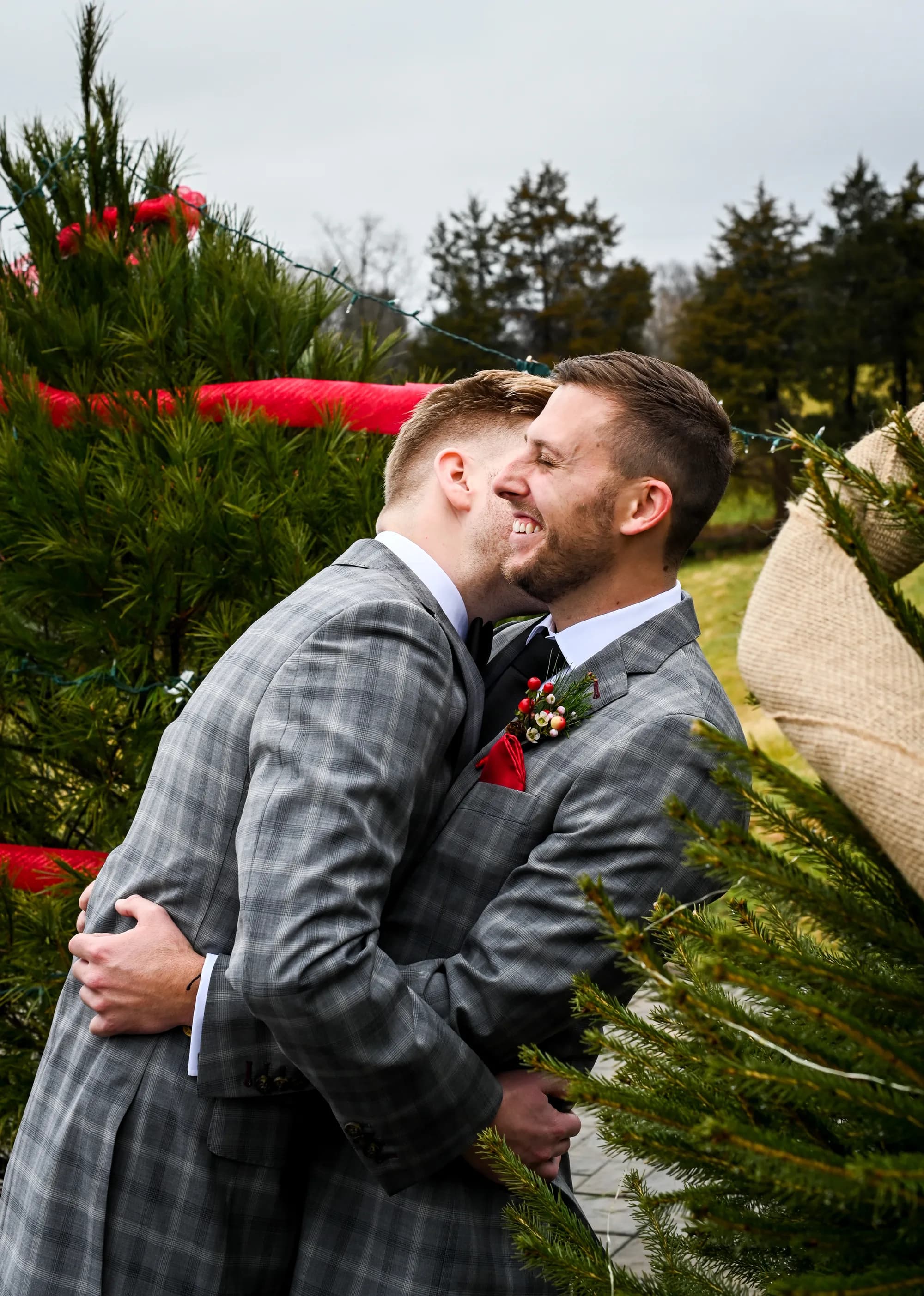 Two grooms in matching gray suits embrace and laugh among Christmas trees at an outdoor winter wedding