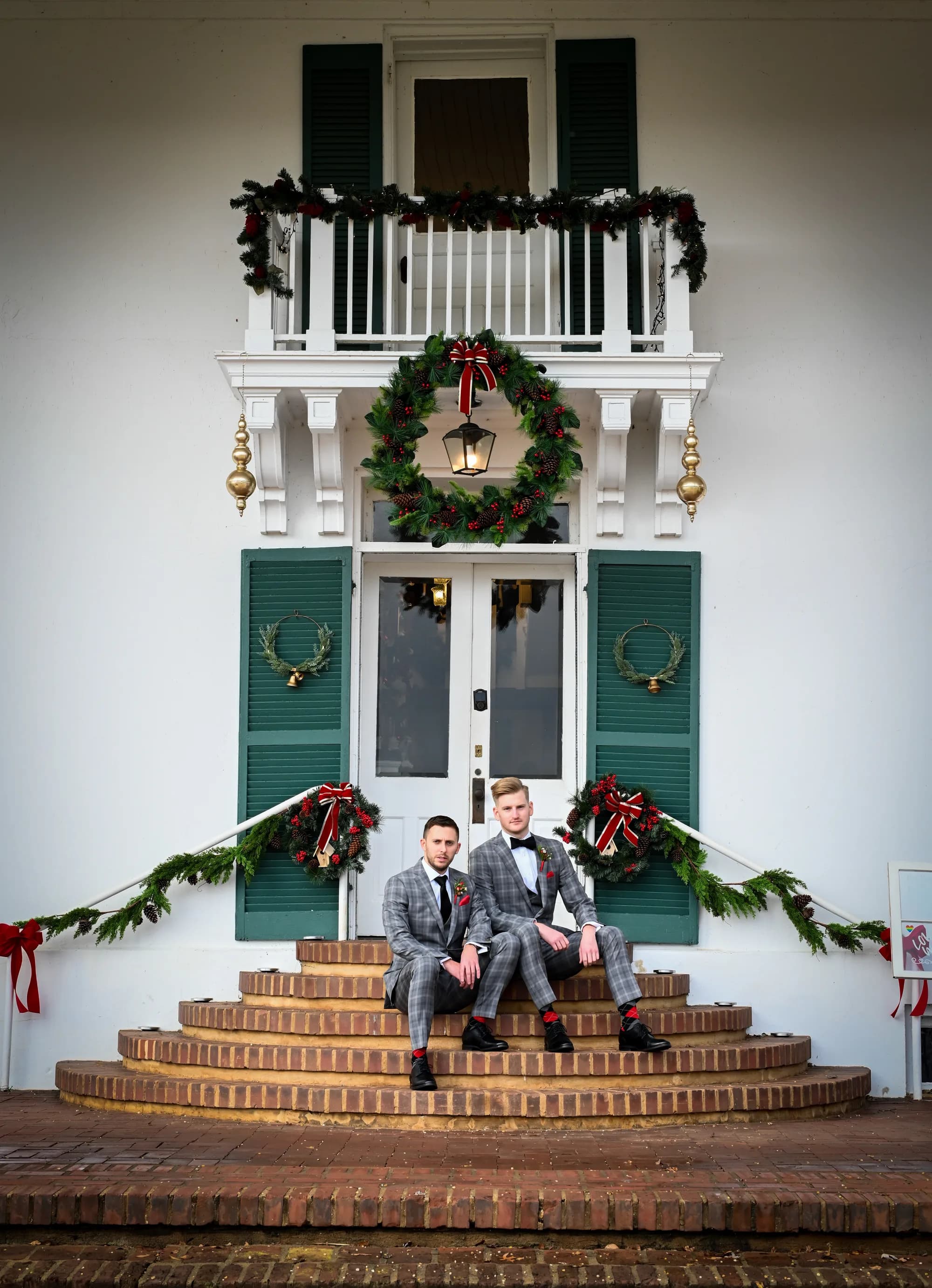 Two grooms in matching plaid suits seated on brick steps of Rixey Manor's Christmas-decorated entrance