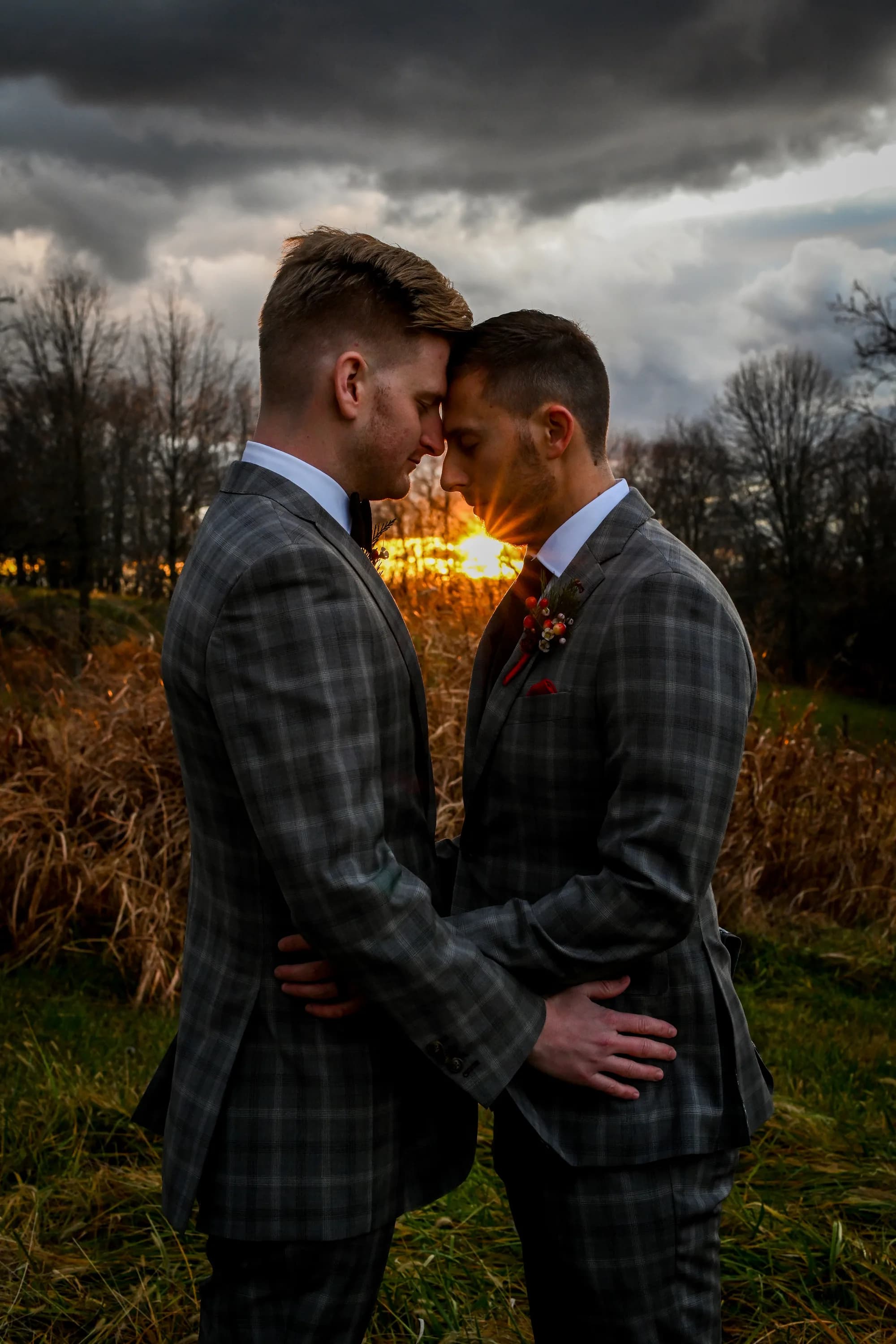 Two grooms in matching plaid suits touch foreheads at golden sunset in a dramatic field setting