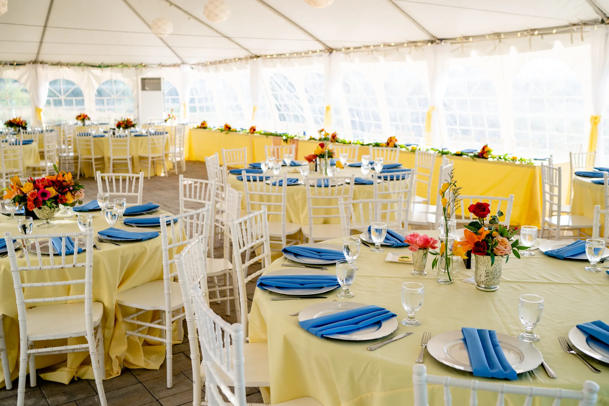 Sunlit reception tent at Rixey Manor with yellow linens, blue napkins, white Chiavari chairs, and colorful floral centerpieces