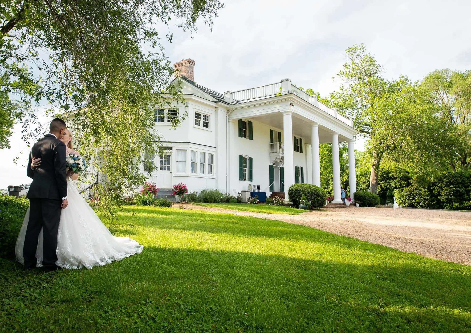 Bride and groom pose on lawn before white columned manor house with trees
