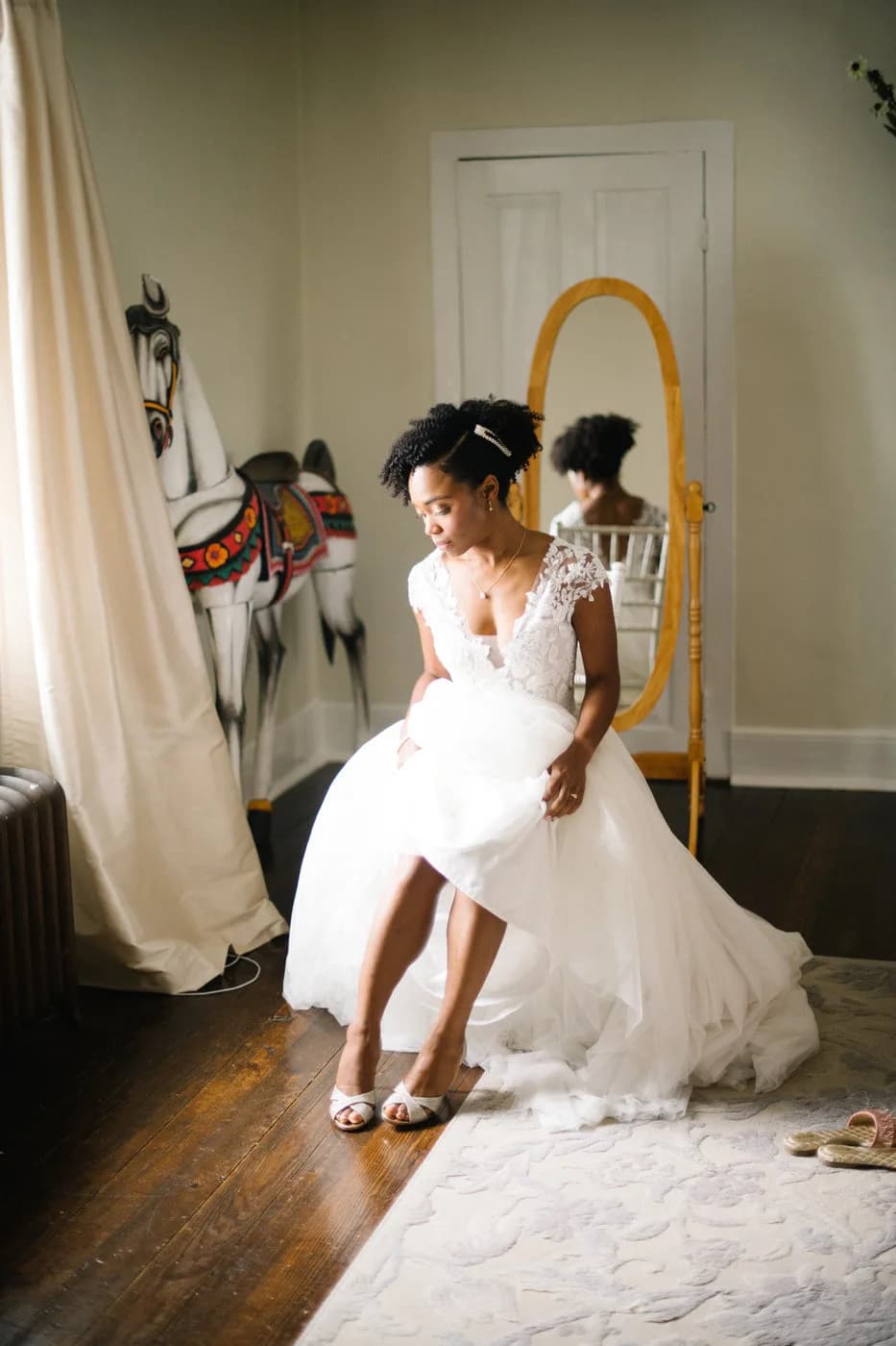Bride in lace wedding gown adjusting her shoes in a warmly lit room with a vintage mirror and carousel horse art
