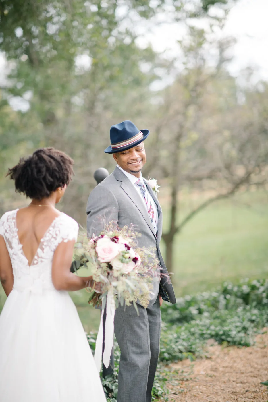 Groom in gray suit and navy hat smiles at bride during first look on wooded estate grounds