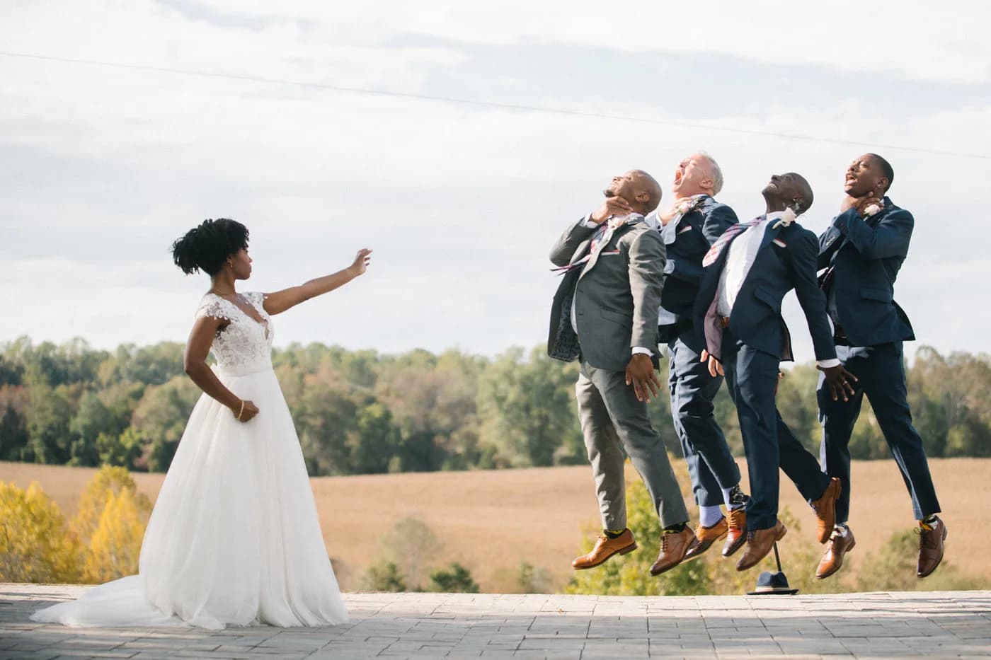 Bride gestures as groomsmen leap into the air on a stone terrace overlooking Virginia countryside