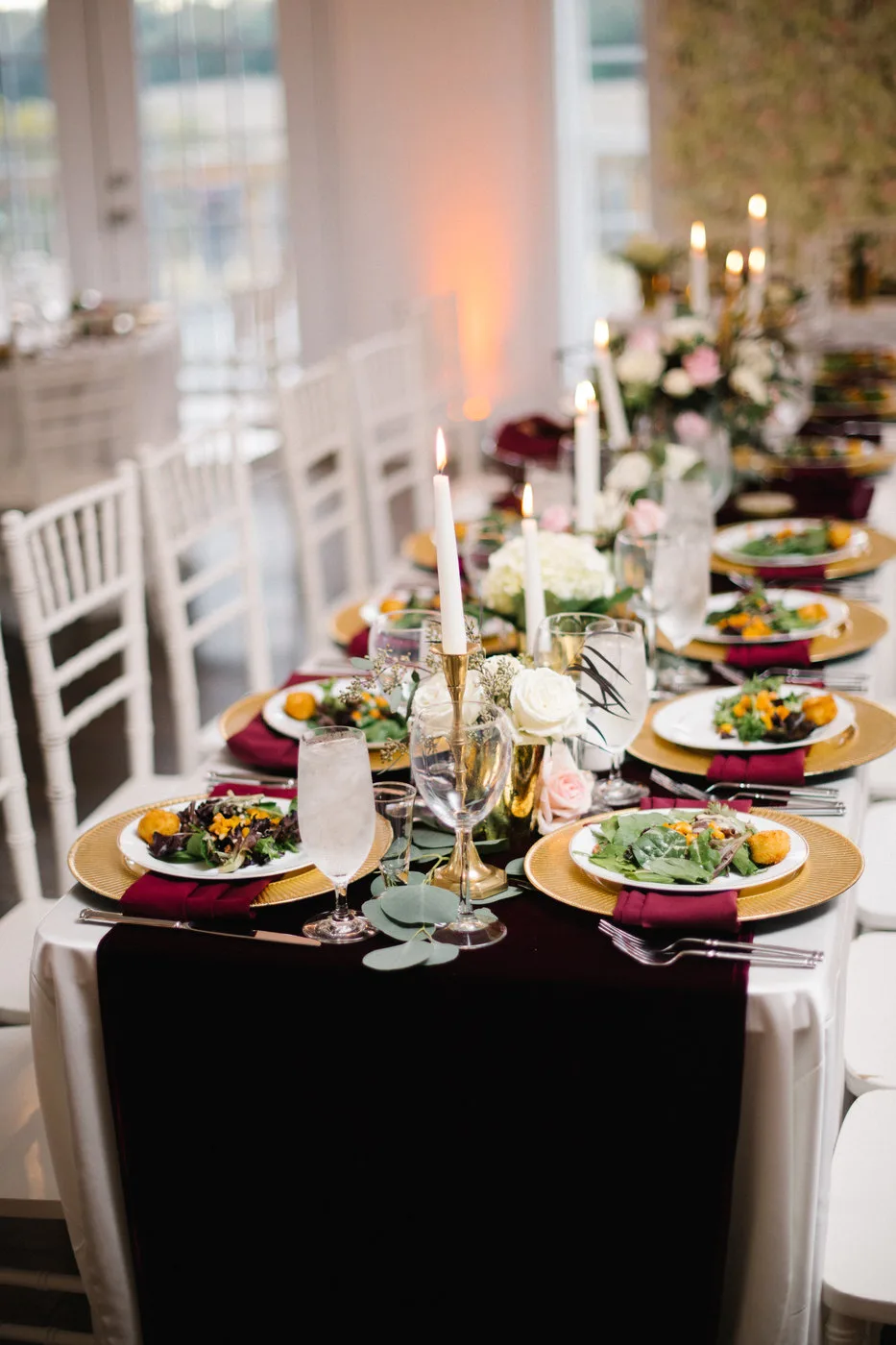 Elegant wedding reception tablescape with gold chargers, burgundy napkins, white candles, and floral centerpieces