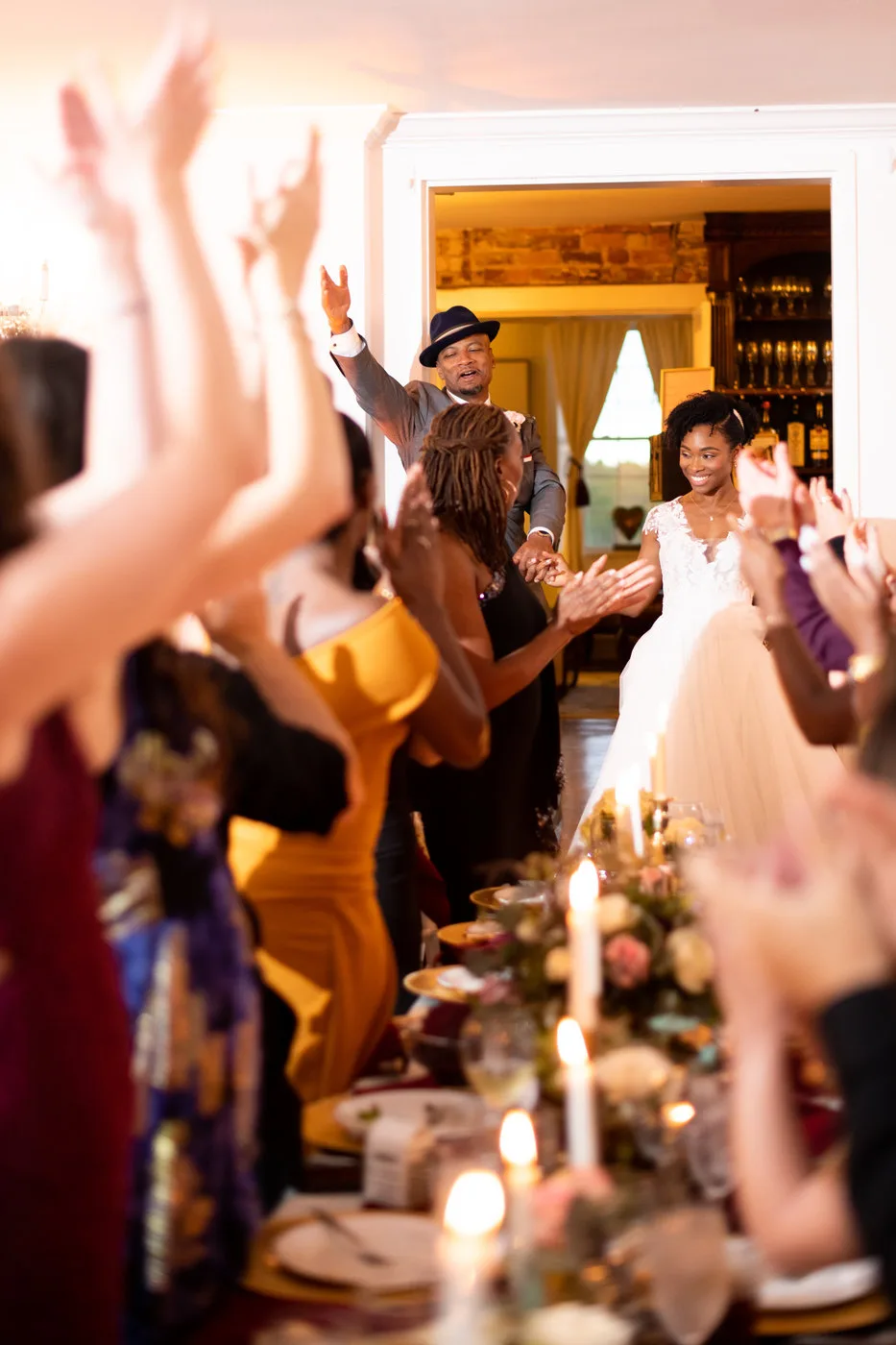 Bride and groom make joyful grand entrance as cheering guests raise hands at candlelit wedding reception
