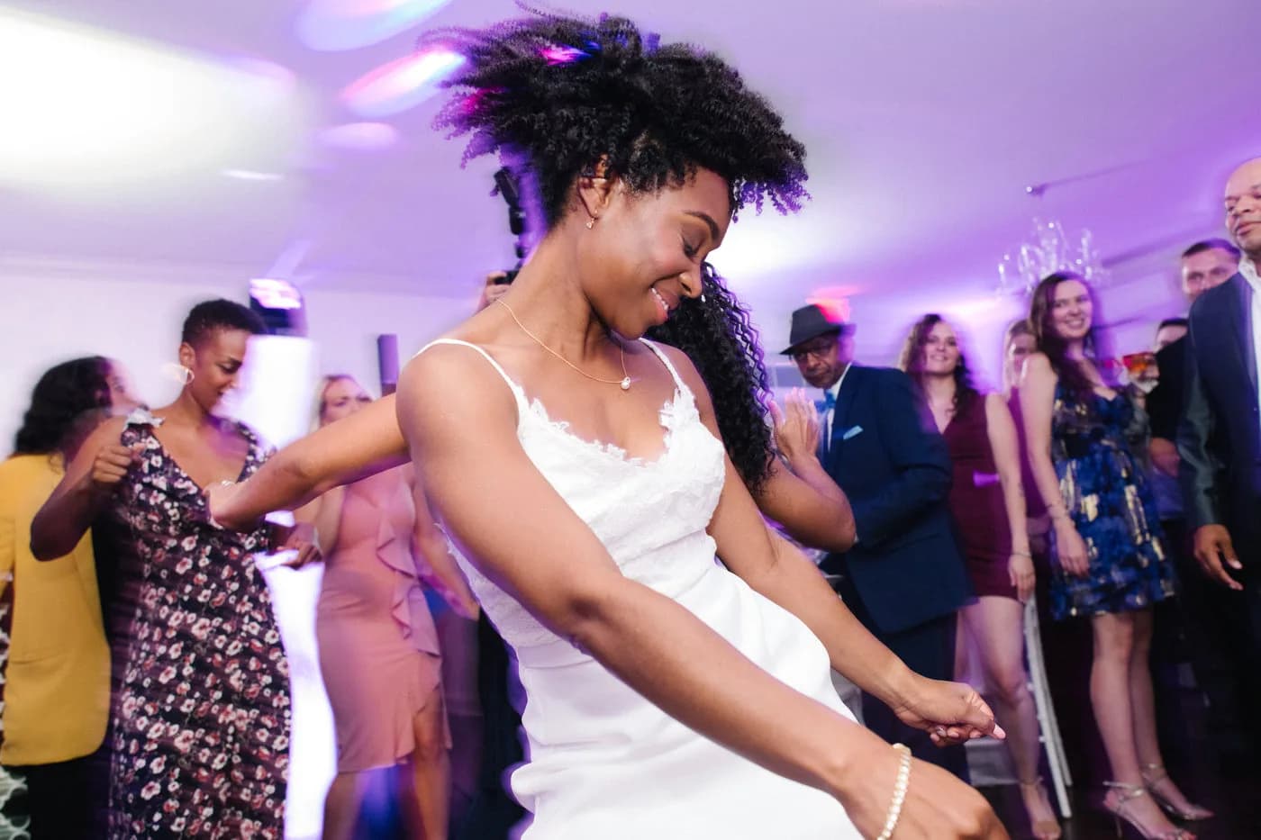 Bride joyfully dancing at wedding reception, hair flying, surrounded by smiling guests under purple lighting