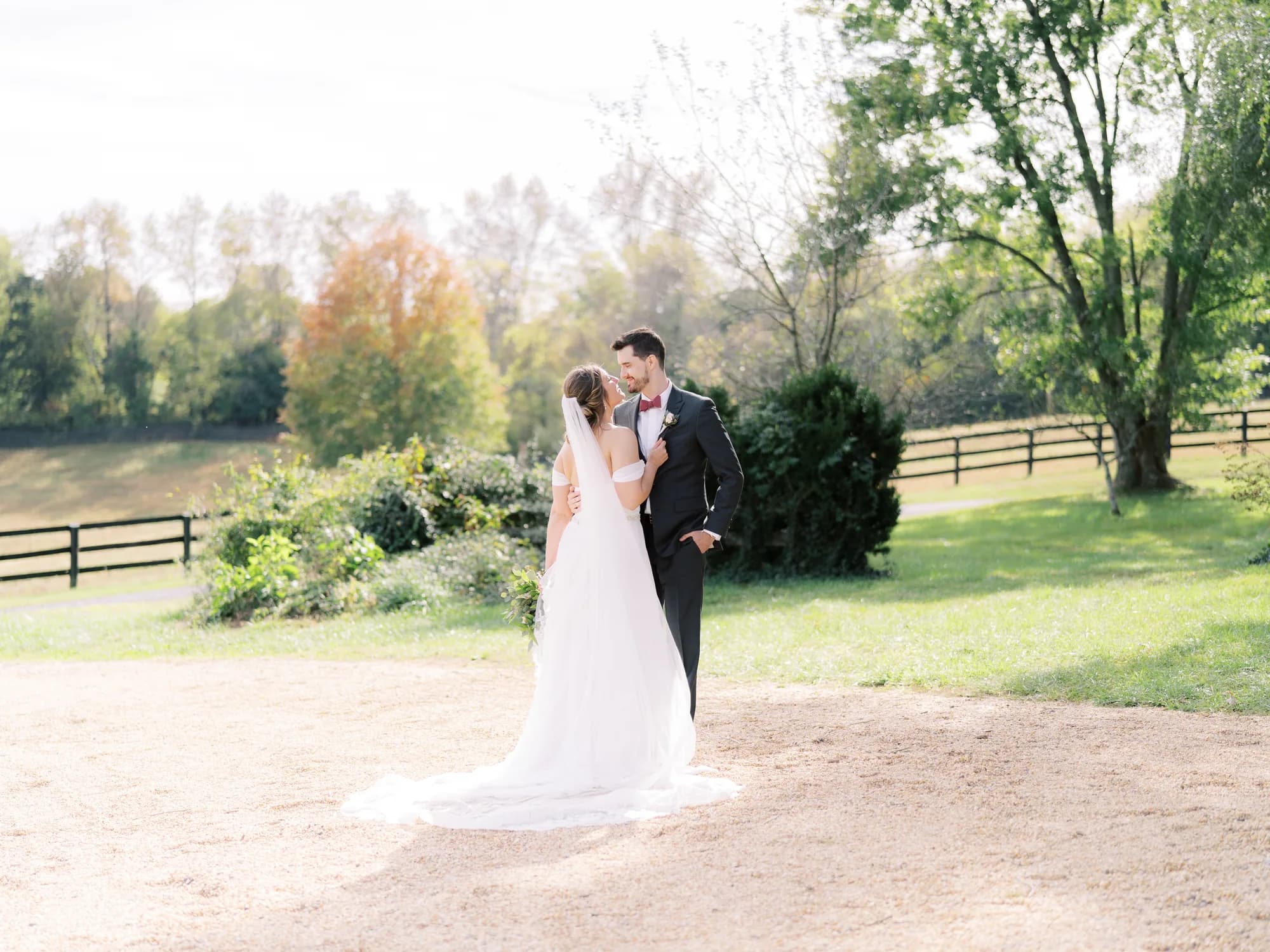 Bride and groom sharing a romantic gaze on sunlit Rixey Manor grounds with lush trees and pastoral fence