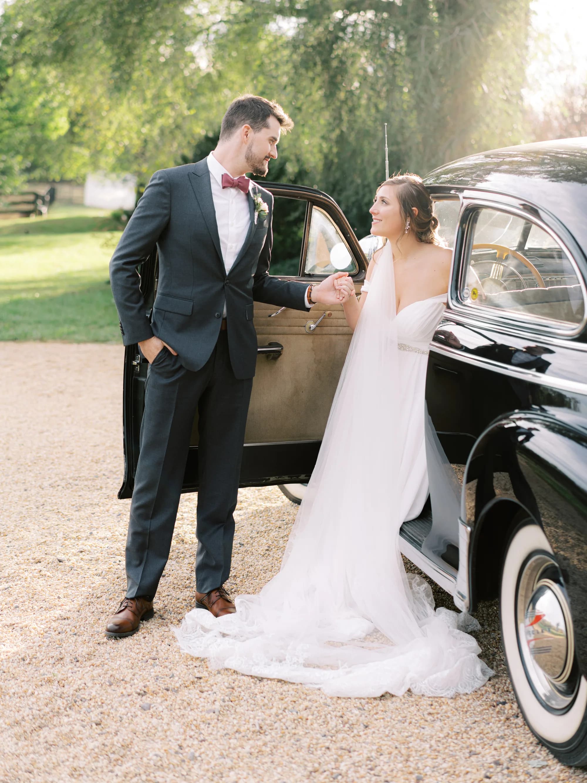 Bride and groom pose beside vintage car with scenic tree-lined background at outdoor venue