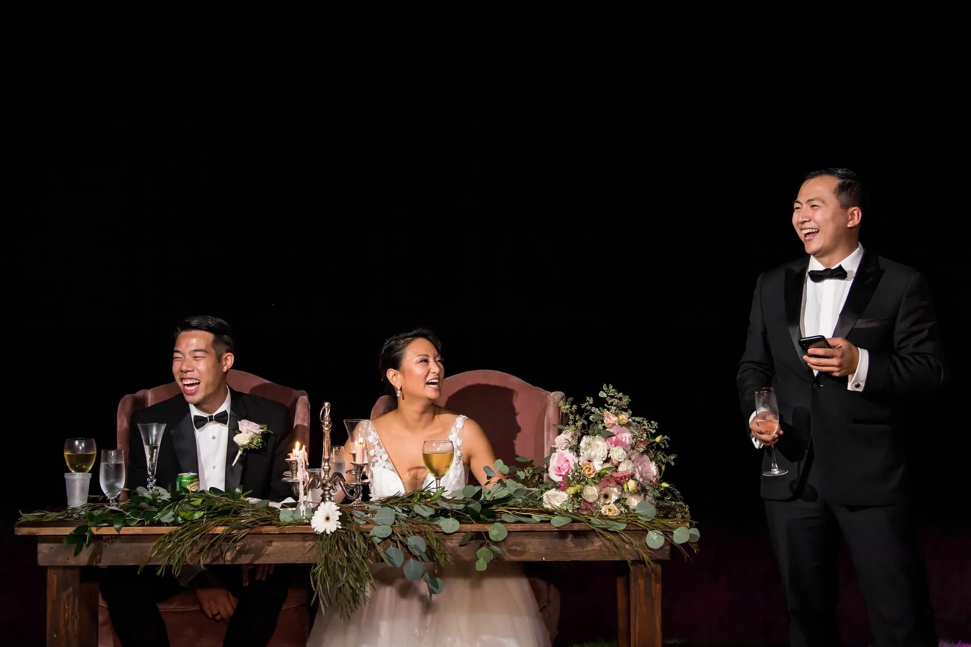 Bride and groom laugh at sweetheart table during a wedding toast, guest speaking with champagne glass at right