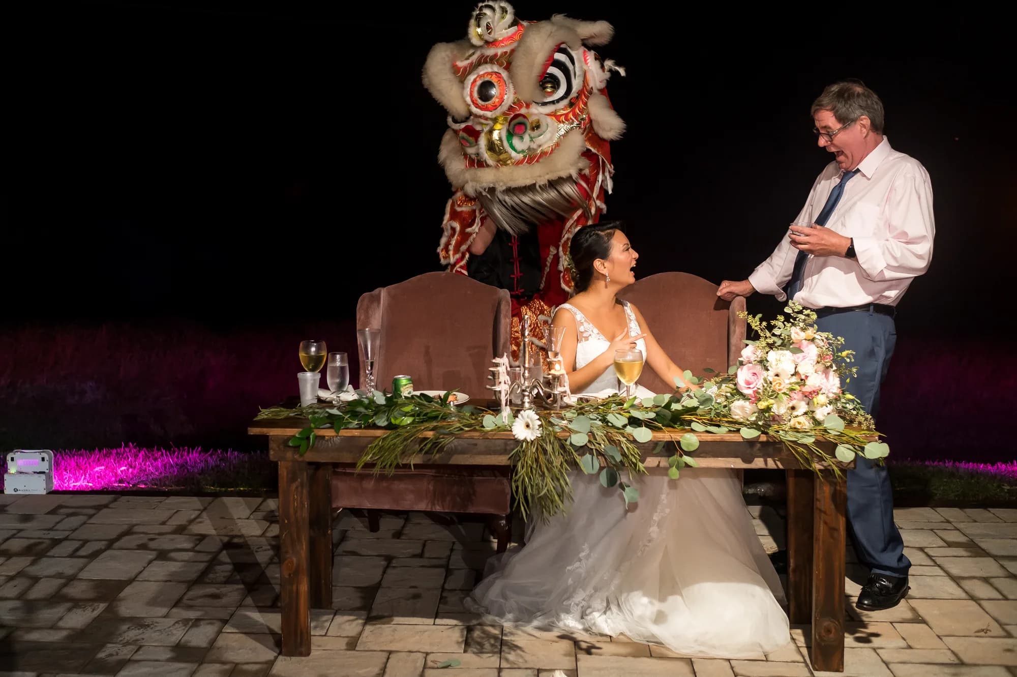 Bride laughing at sweetheart table as Chinese lion dancer performs behind her at outdoor wedding reception