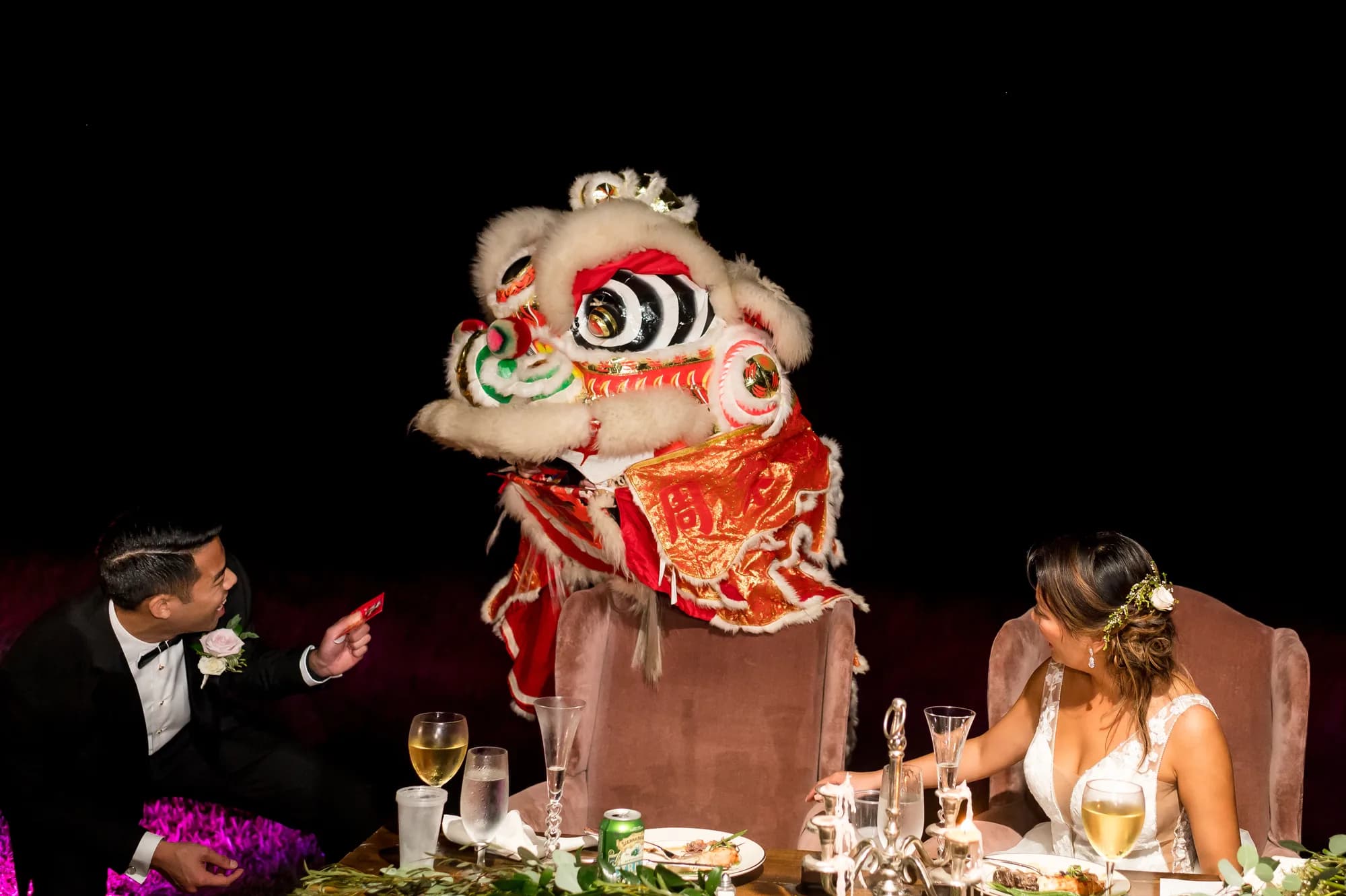 Bride and groom delight as a traditional Chinese lion dance performer visits their sweetheart table at wedding reception
