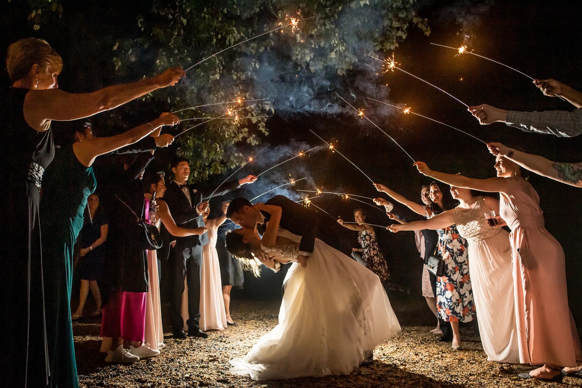 Bride and groom share a dip kiss under an arch of sparklers held by cheering wedding guests at night