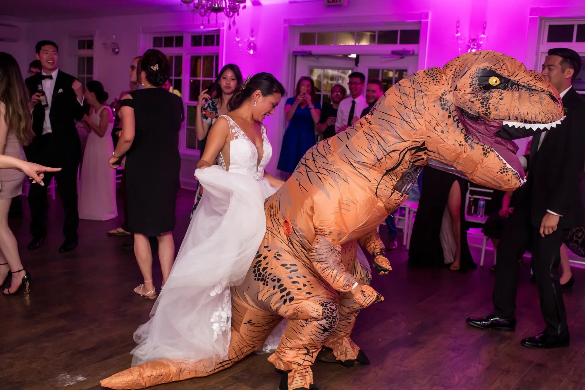 Bride laughing while dancing with a guest in an inflatable T-Rex costume at a lively wedding reception