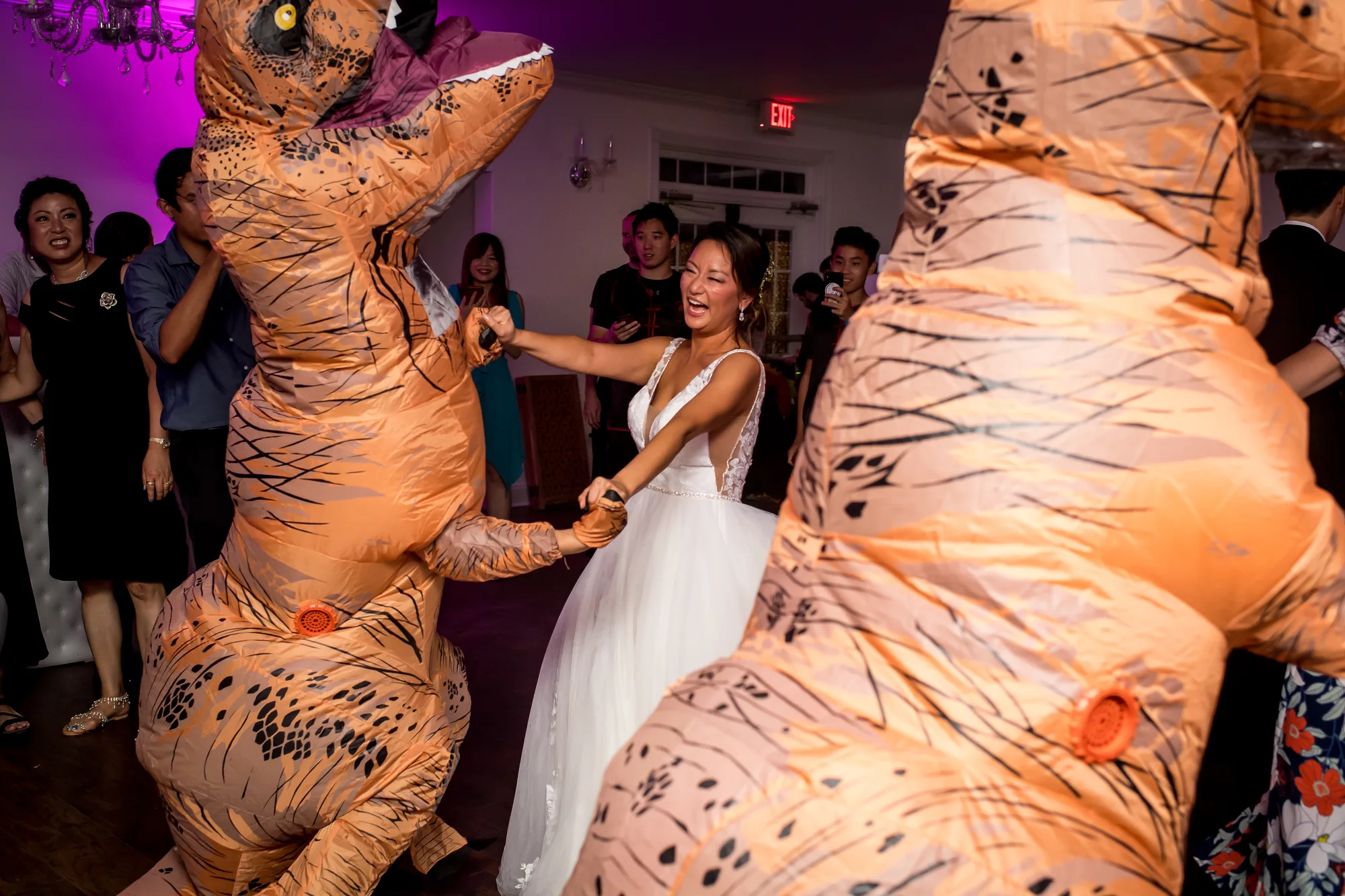 Laughing bride dances with guests in inflatable T-Rex costumes at lively wedding reception