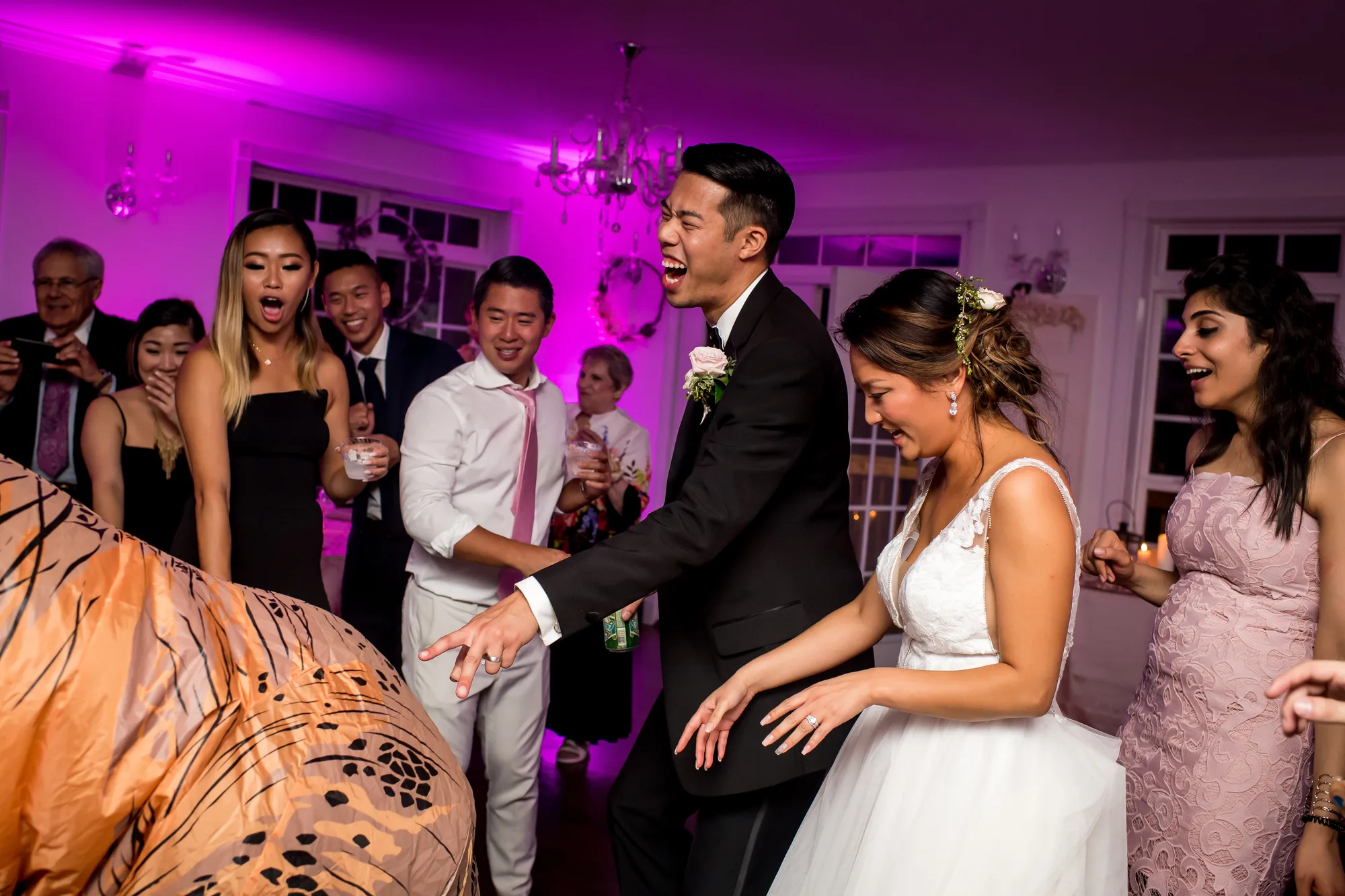 Bride and groom dancing at wedding reception with guests in magenta-lit venue, man in tuxedo and woman in white dress