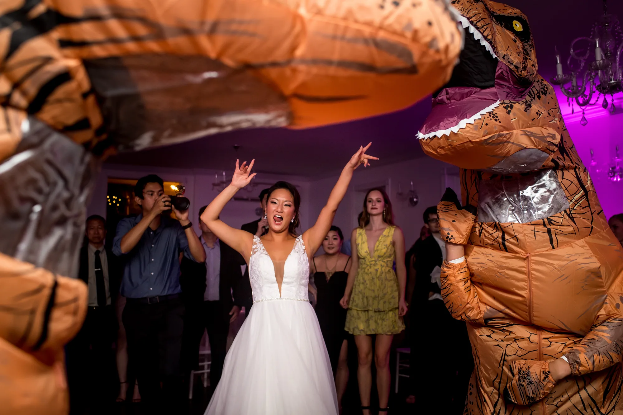 Bride laughs with arms raised between two inflatable T-Rex costumes on the wedding reception dance floor