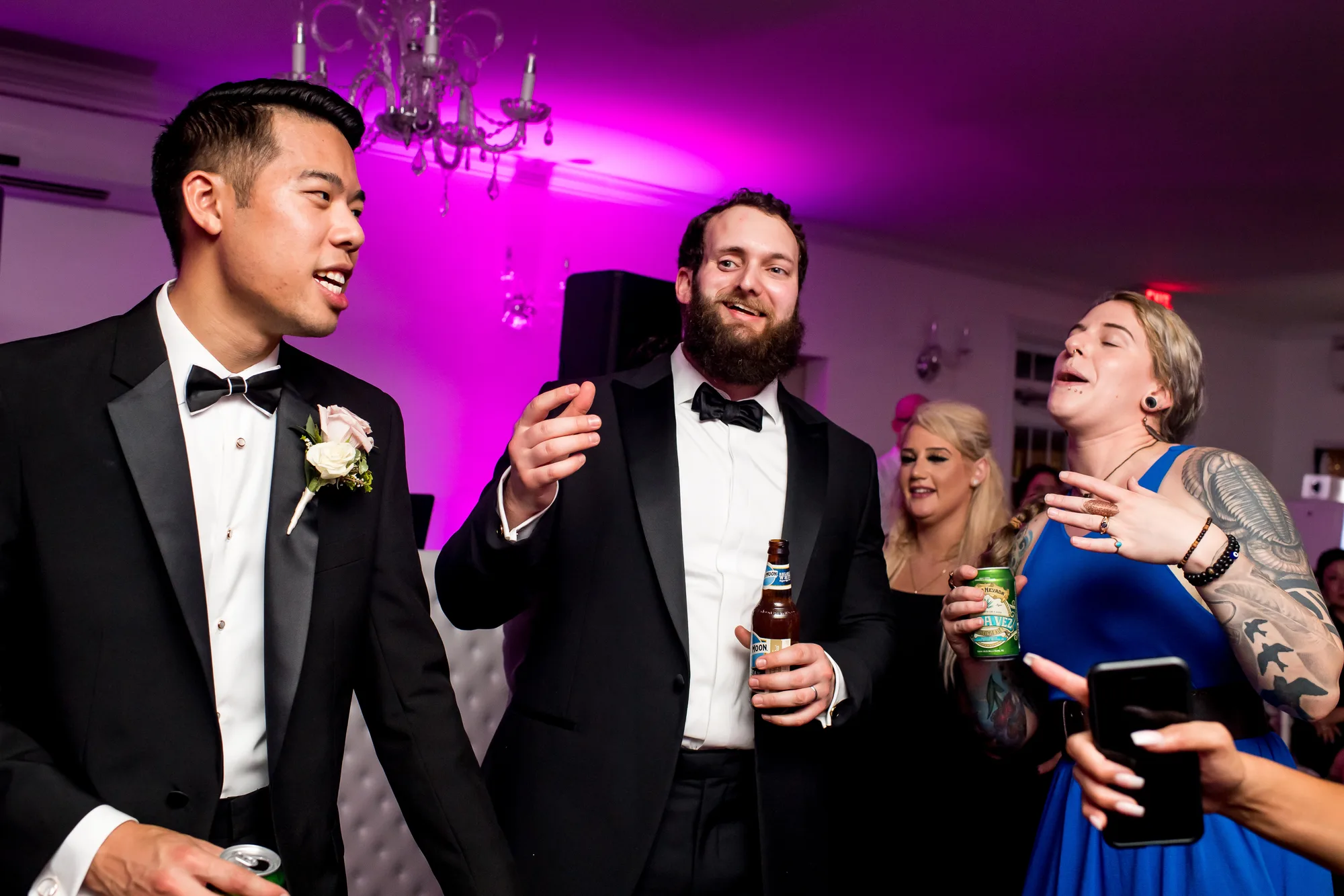 Groom and groomsmen in tuxedos celebrate at reception with purple ambient lighting and guests in background