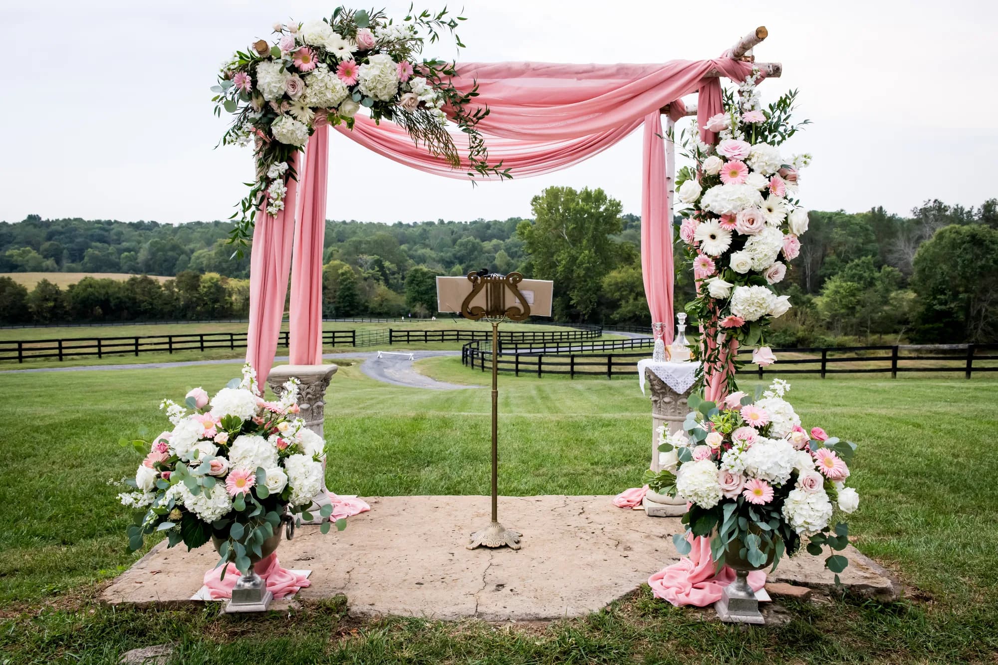 Pink draped floral arch with white and blush blooms on Rixey Manor's green lawn overlooking fenced countryside