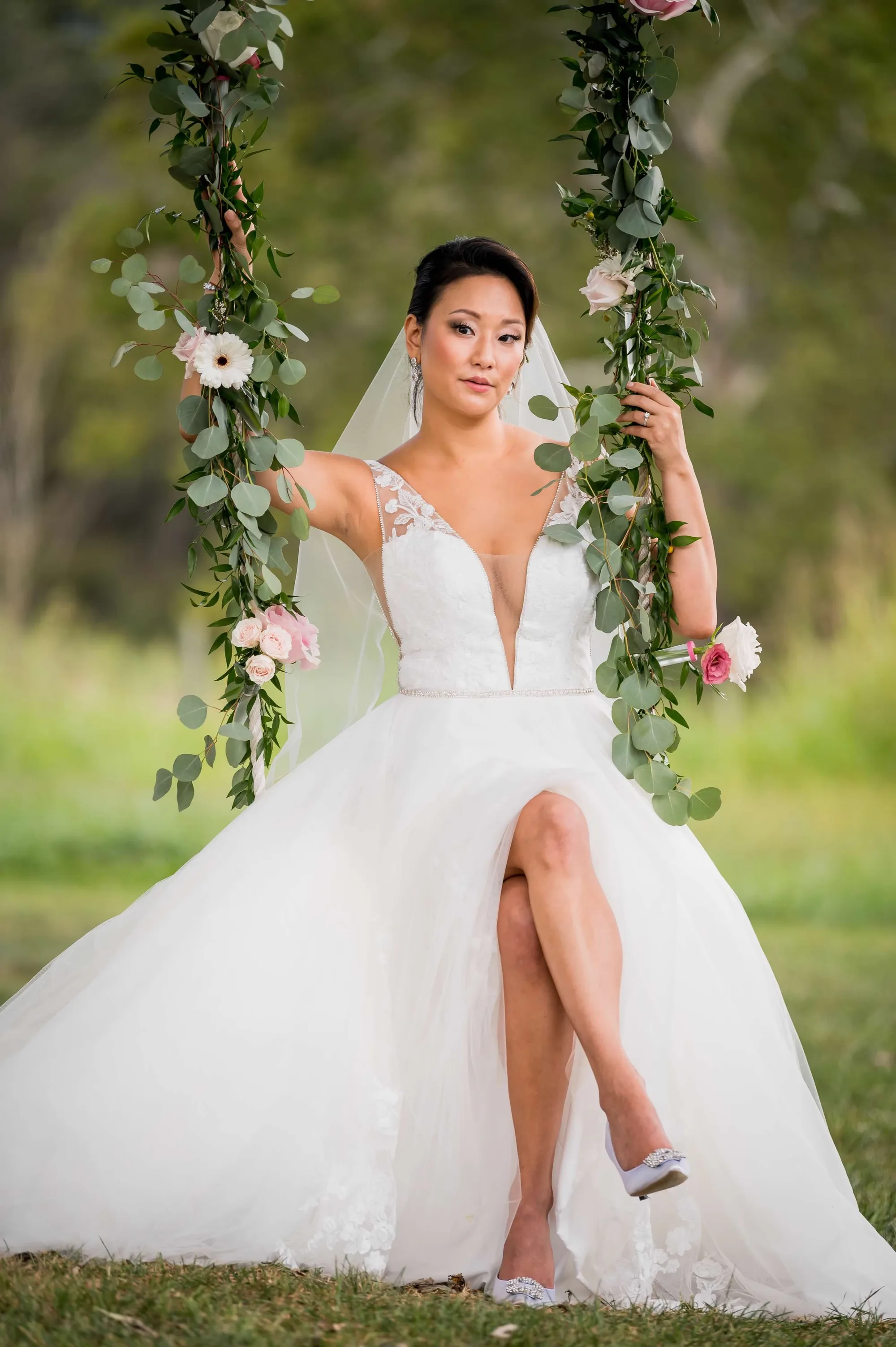 Bride in deep-V ballgown seated on floral swing adorned with eucalyptus and pink roses in lush outdoor setting