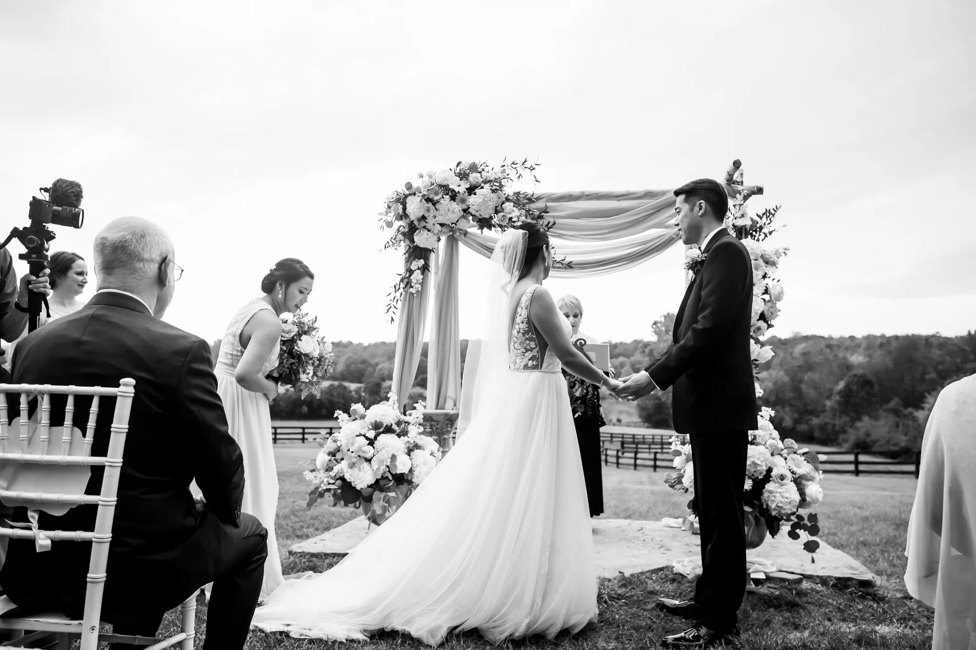 Black and white wedding ceremony photo showing bride and groom at altar with floral arch in rural Virginia field.