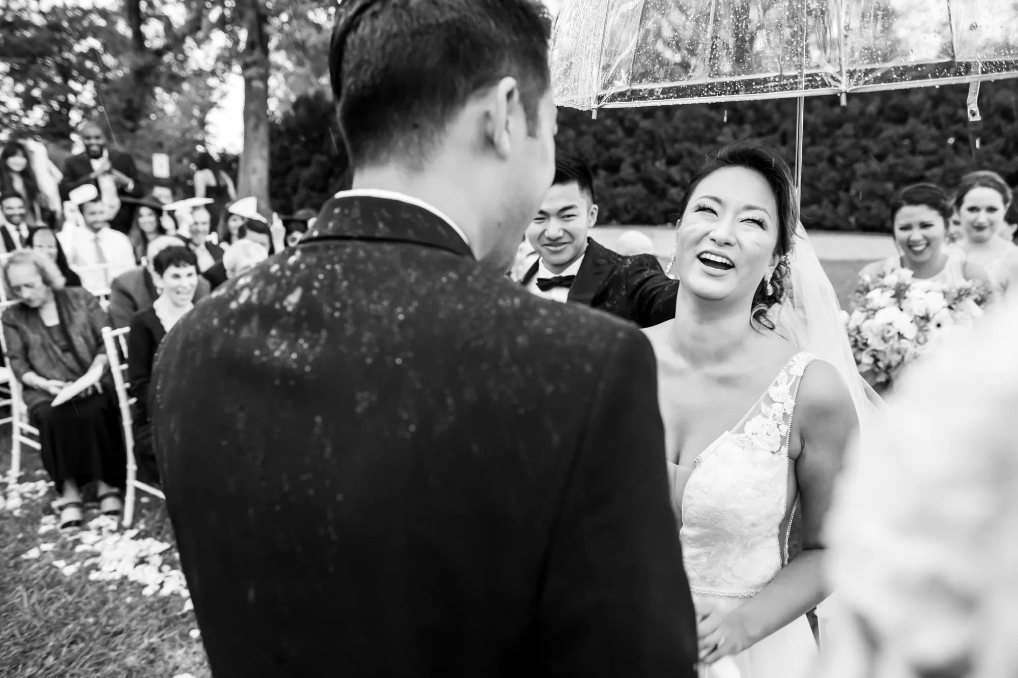 Bride laughs joyfully under clear umbrella during rainy outdoor wedding ceremony as guests smile behind her