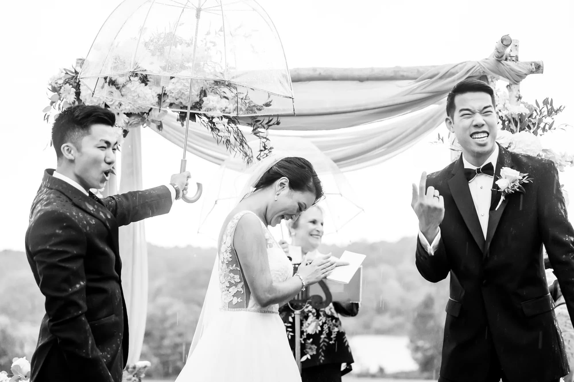Bride laughs while reading vows as groom and groomsman react with joy during rainy outdoor ceremony