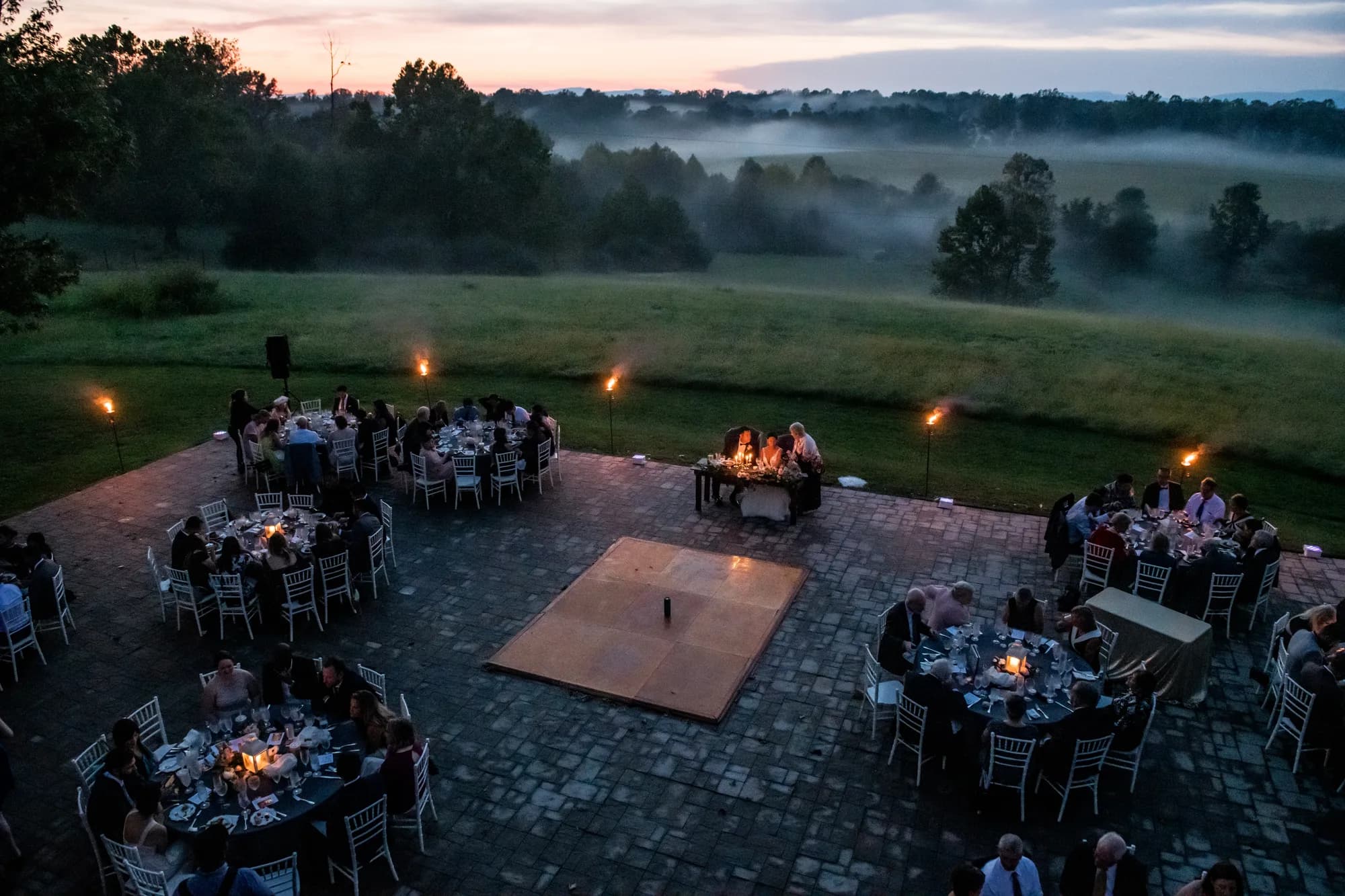 Aerial dusk view of outdoor wedding reception on stone terrace with torches and misty Virginia countryside