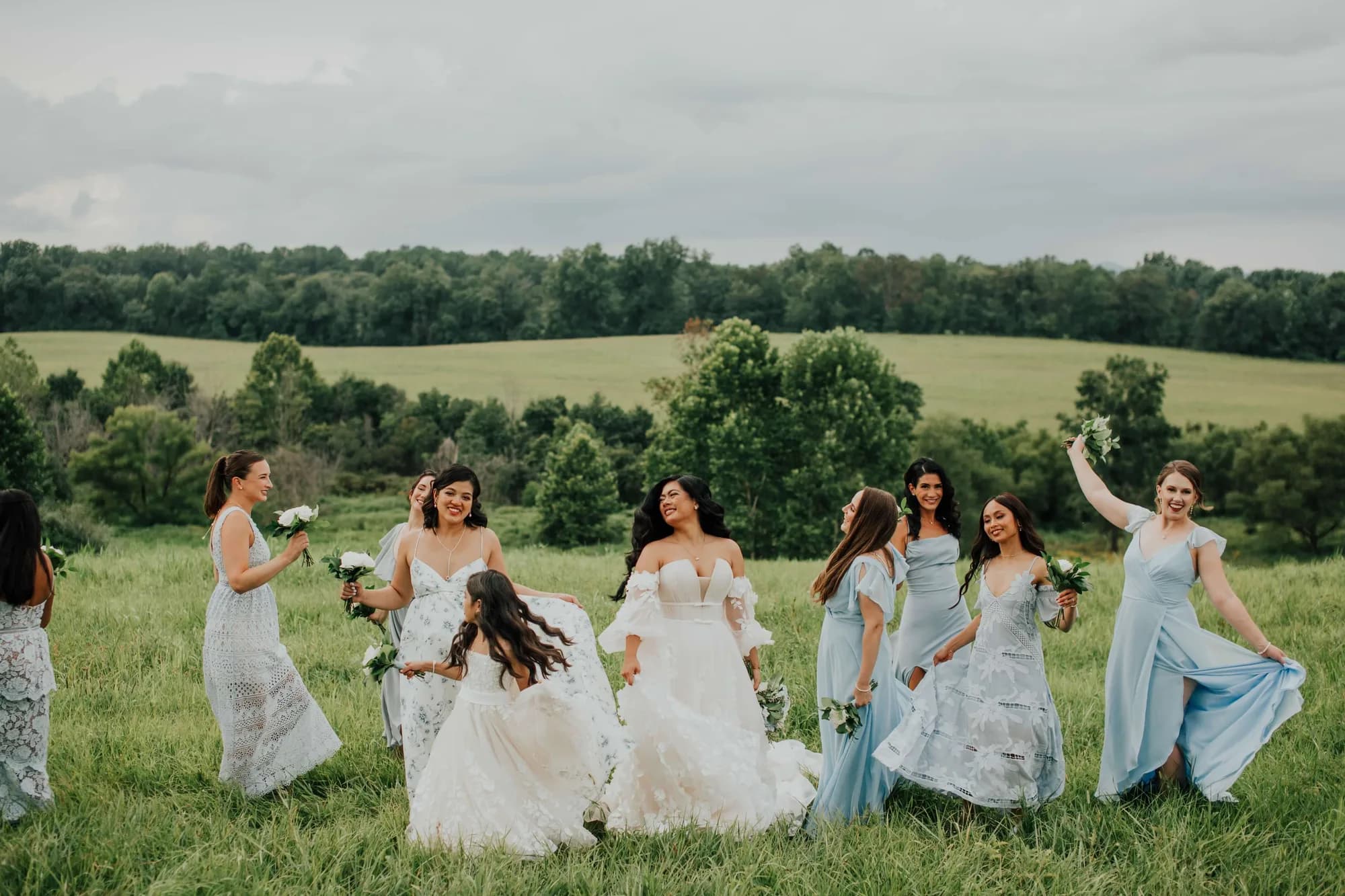 Bride and bridesmaids in blue dresses laugh and dance in lush green Virginia countryside fields