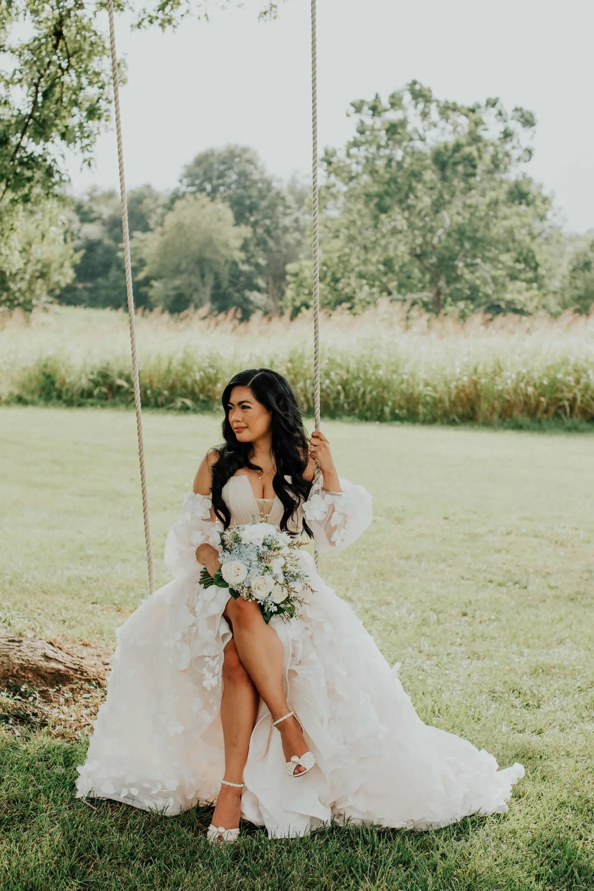 Bride in ruffled gown holding white bouquet seated on rope swing amid lush Virginia countryside grounds