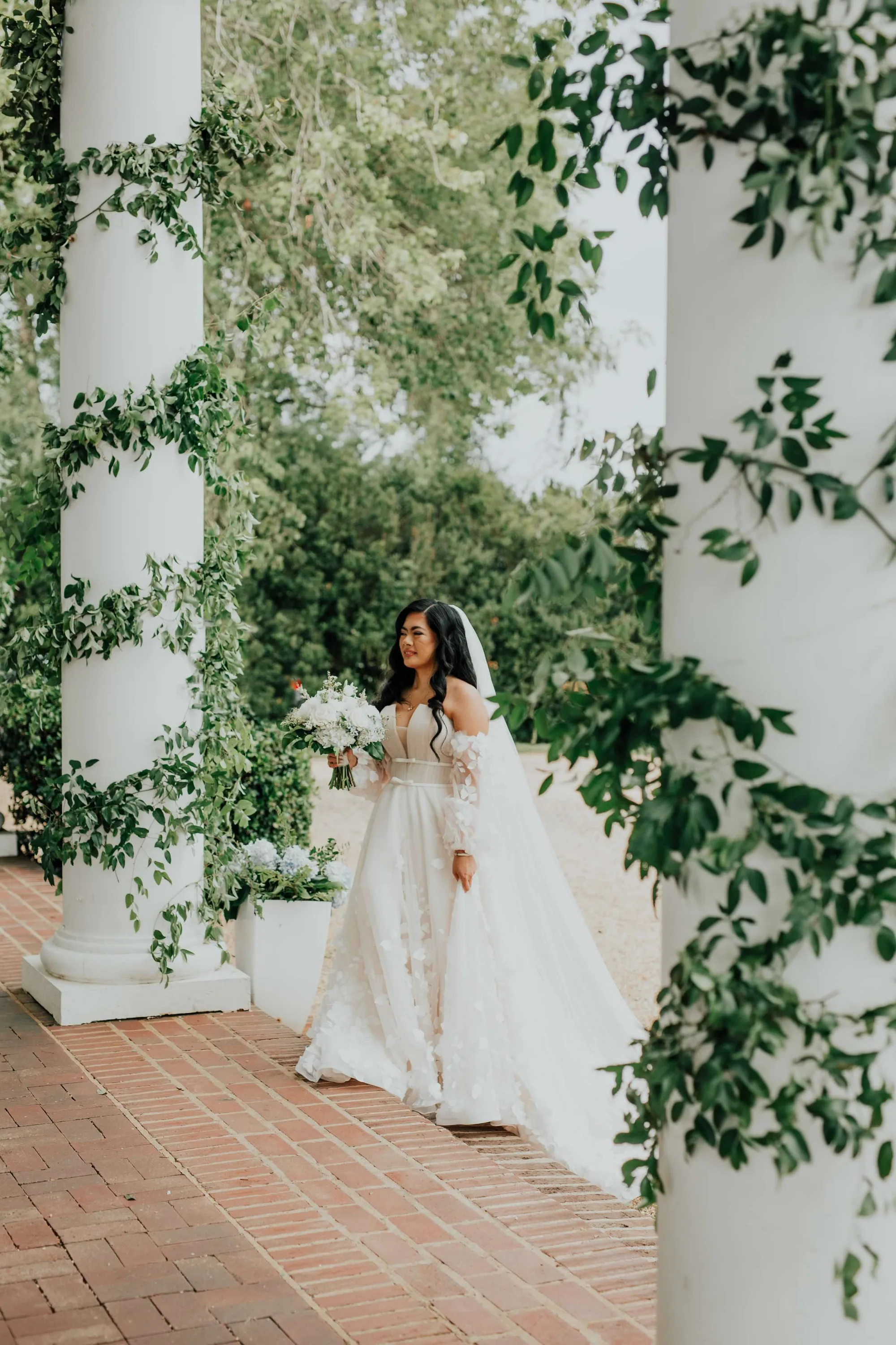 Bride in flowing gown walks among ivy-draped white columns on brick colonnade at Rixey Manor