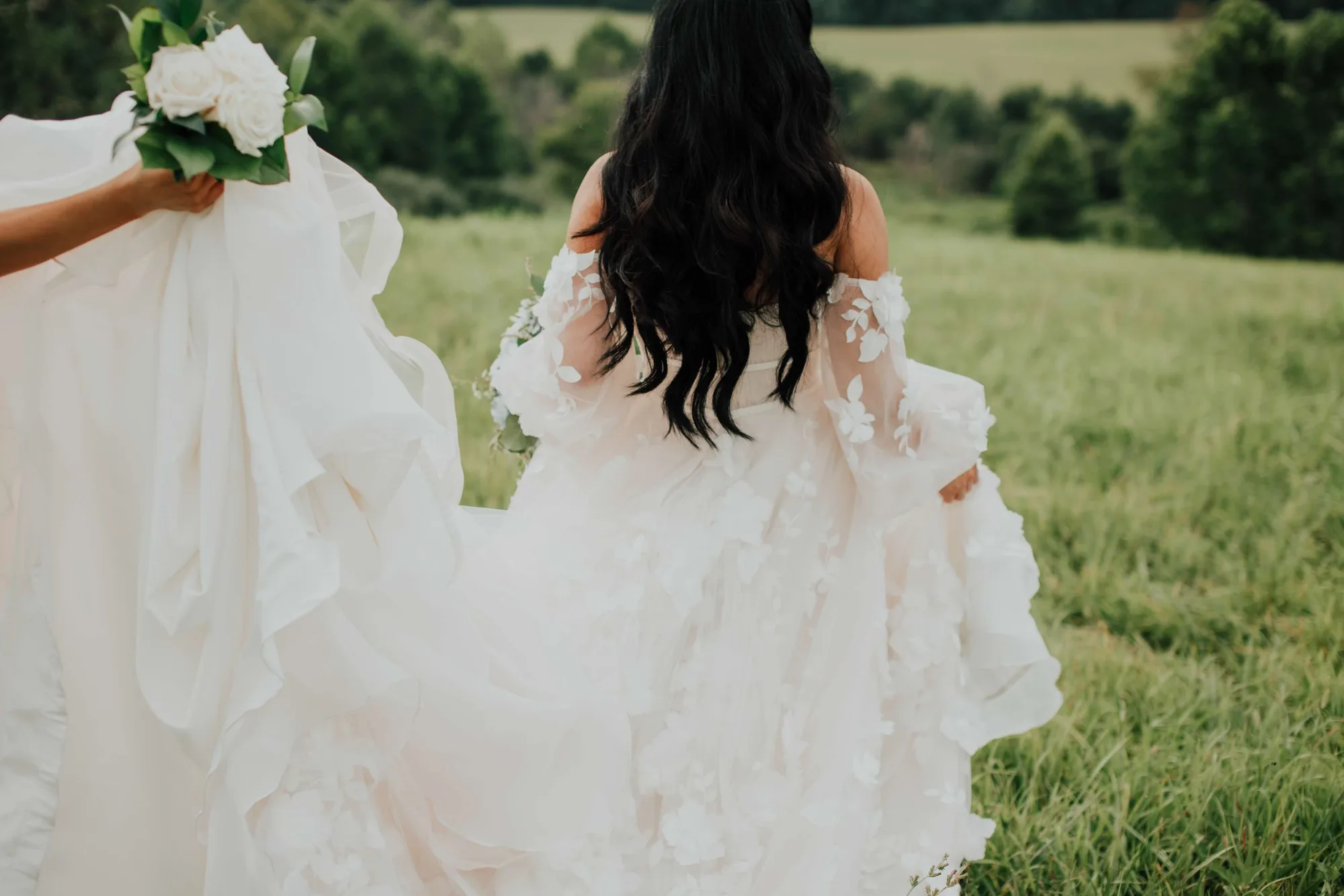 Bride in floral lace gown walks through lush green Virginia countryside fields, train carried behind her