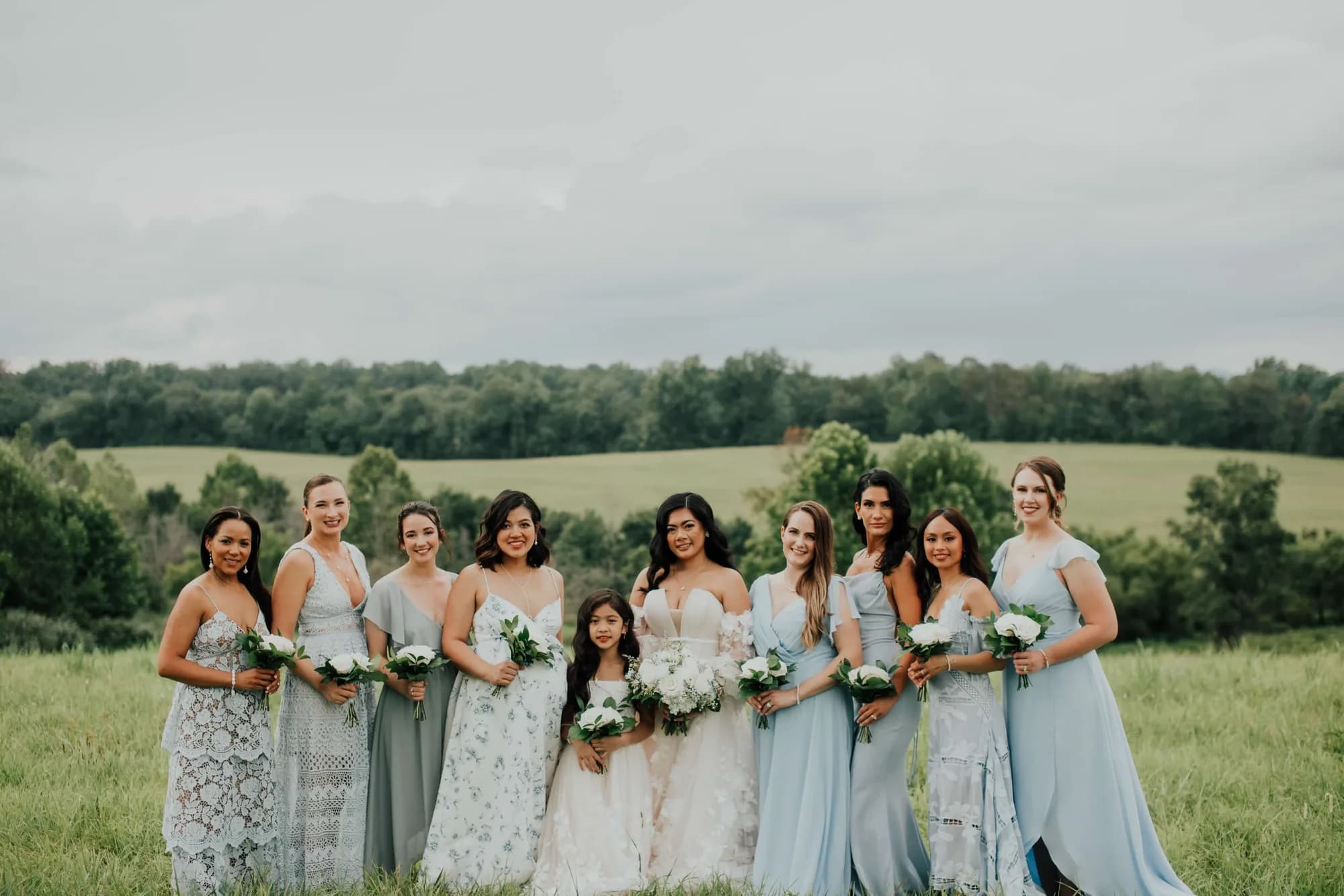 Bride with bridesmaids in sage and blue gowns posed in lush green Virginia countryside at Rixey Manor