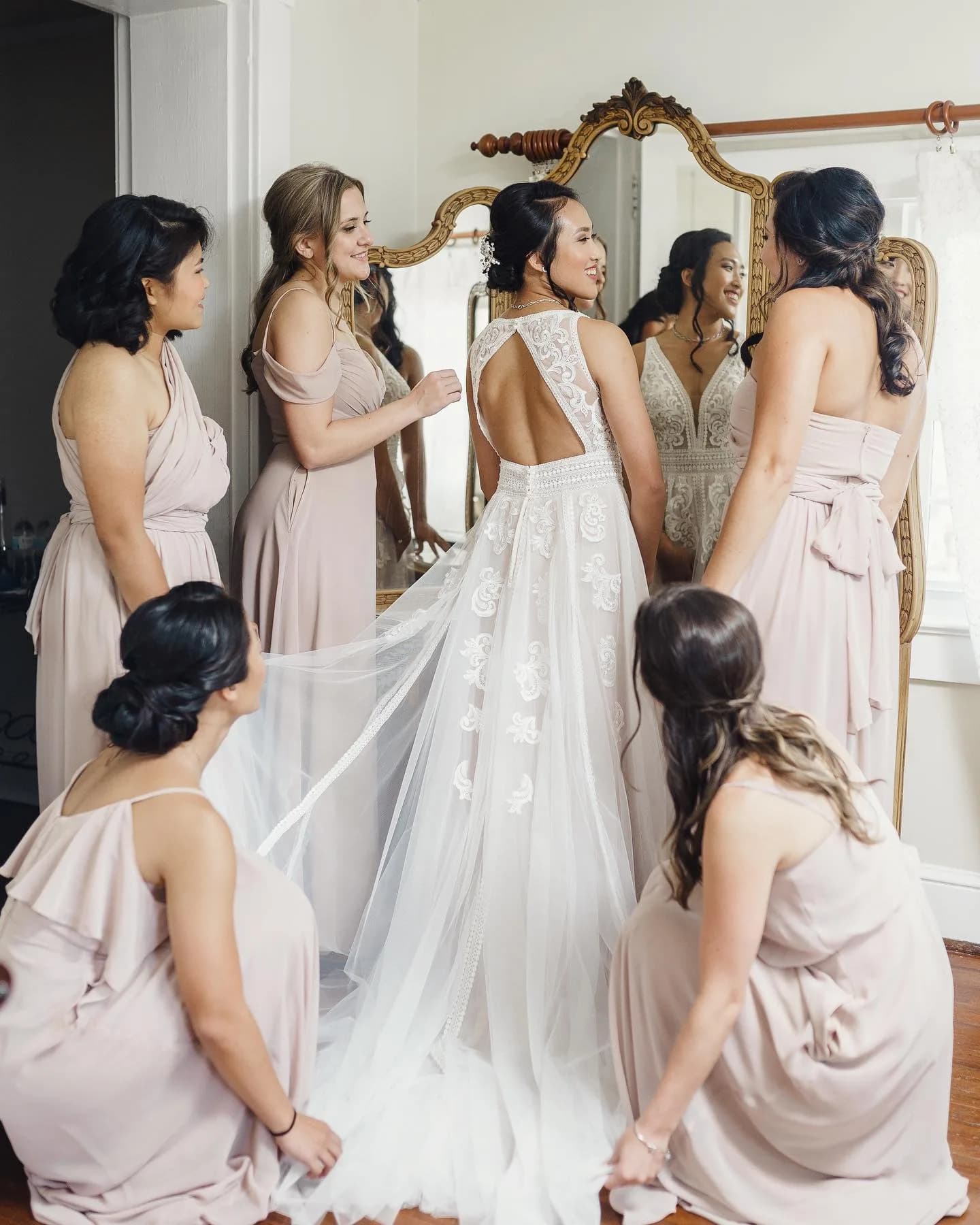 Bride in lace gown surrounded by bridesmaids in blush dresses before a gilded mirror during getting-ready moment