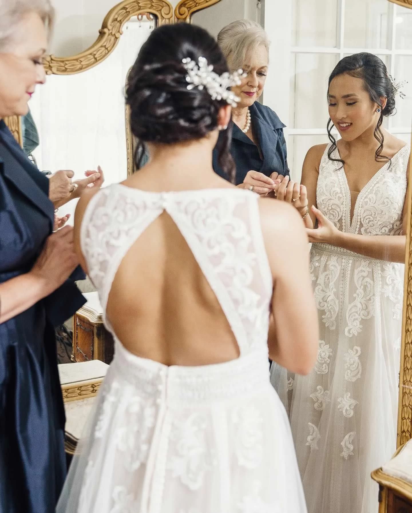 Bride in open-back lace gown assisted by bridesmaids and family before her wedding ceremony