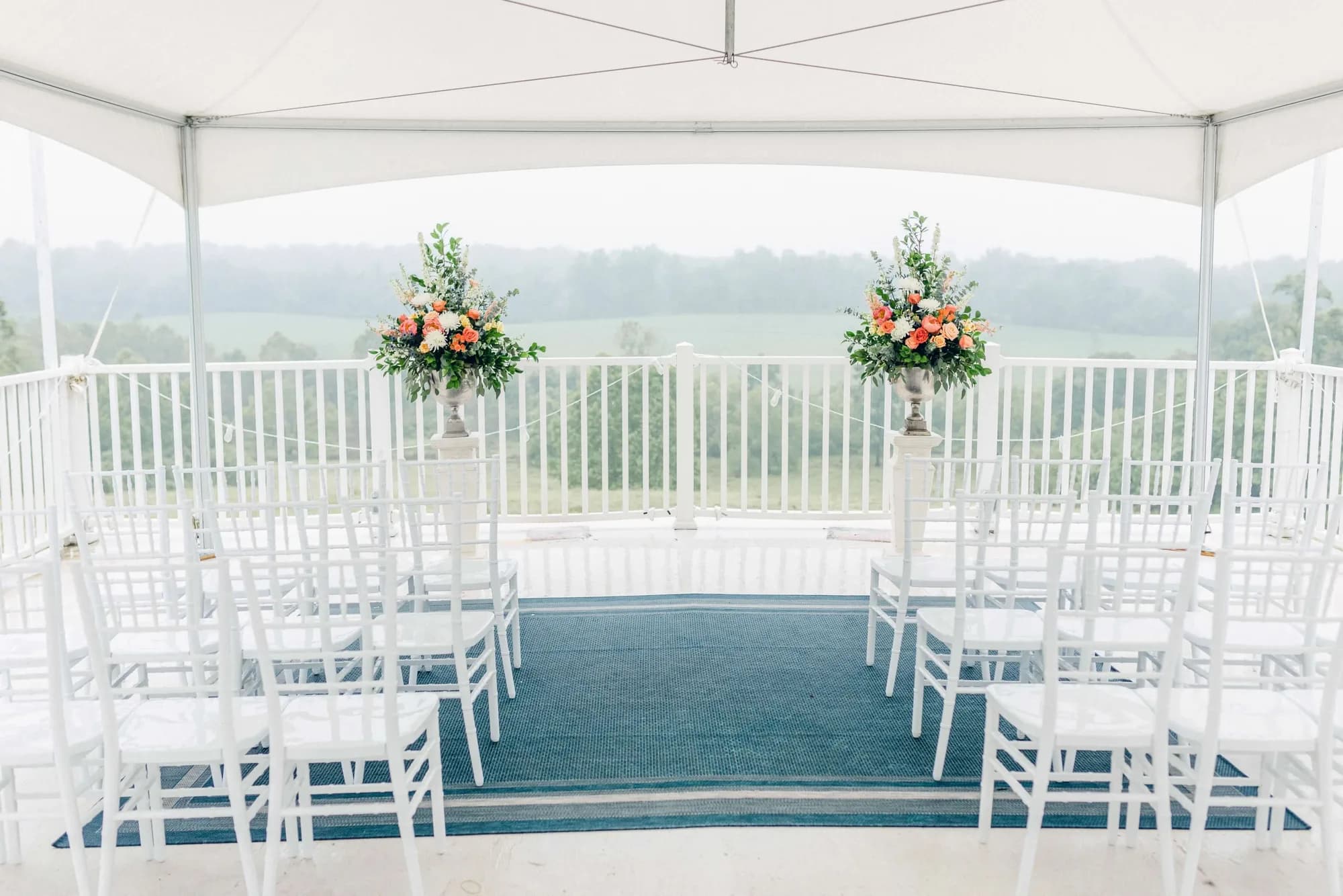White chiavari chairs and floral arrangements set for a wedding ceremony under a tent at Rixey Manor with misty rural views