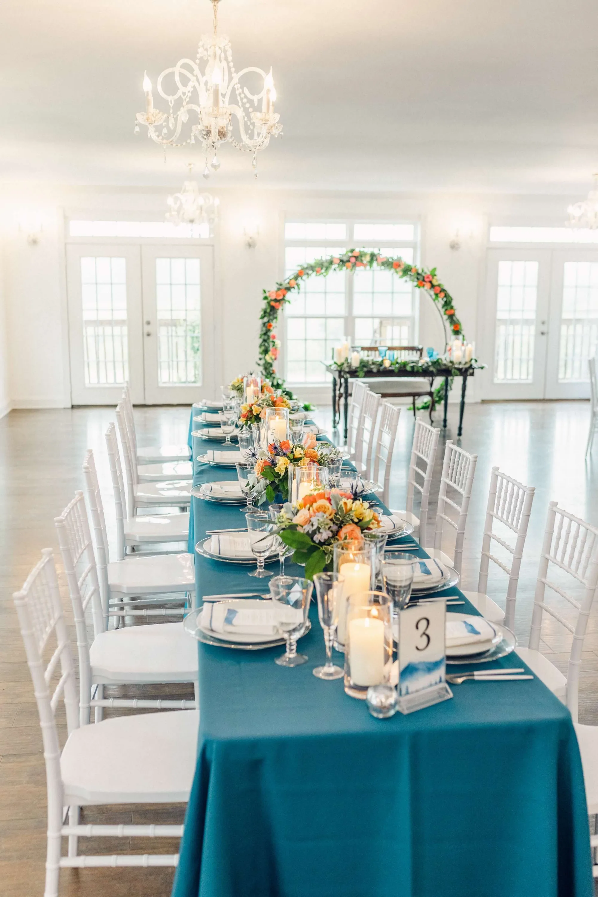 Elegant wedding reception with teal tablecloth, orange floral arrangements, white chiavari chairs, and decorative arch