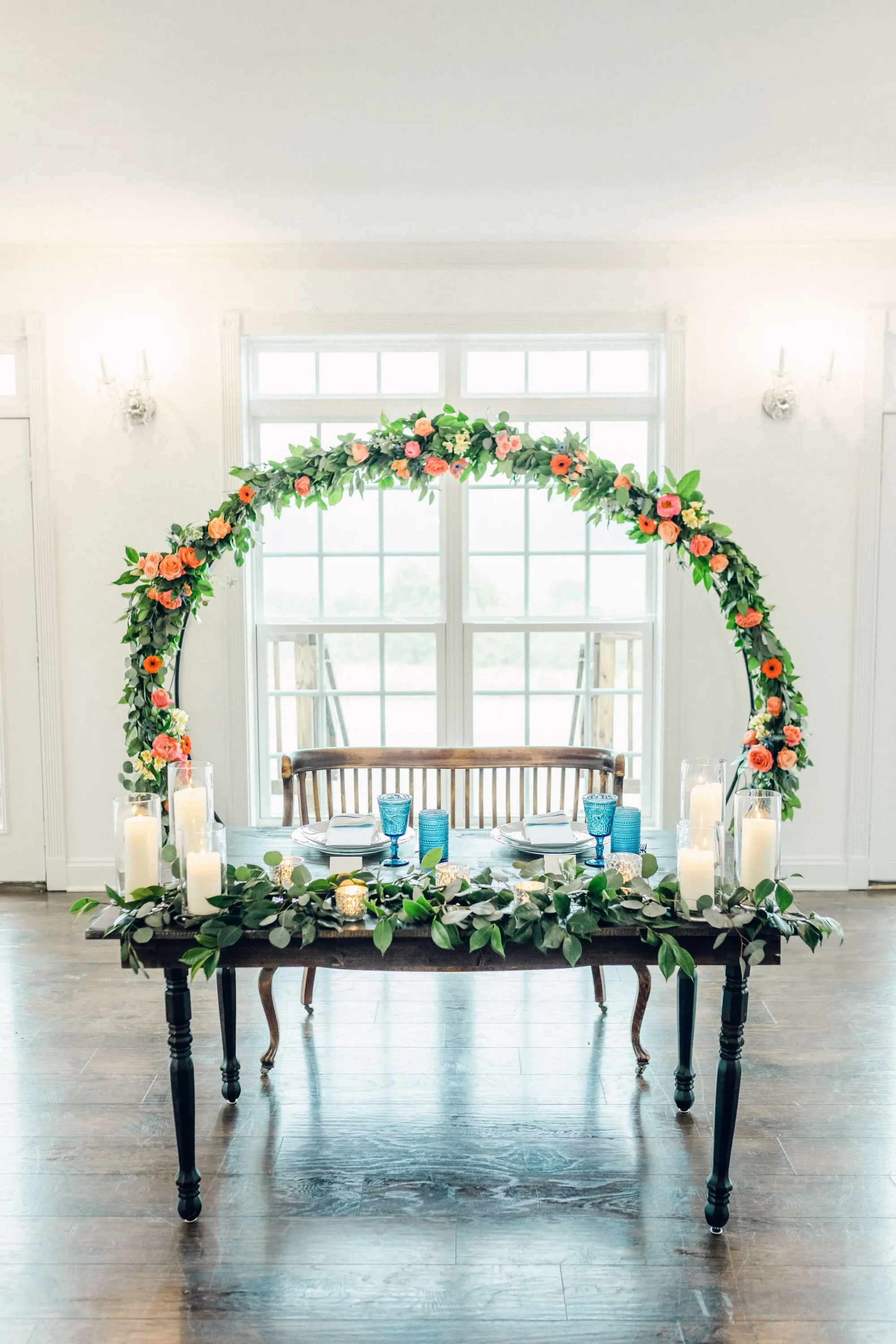 Sweetheart table with floral arch, candles, and blue goblets in a bright white Rixey Manor reception room