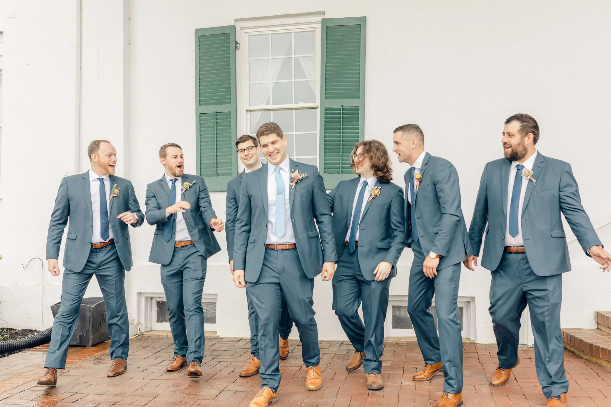 Groom and groomsmen laughing and walking together in navy suits outside a white colonial building