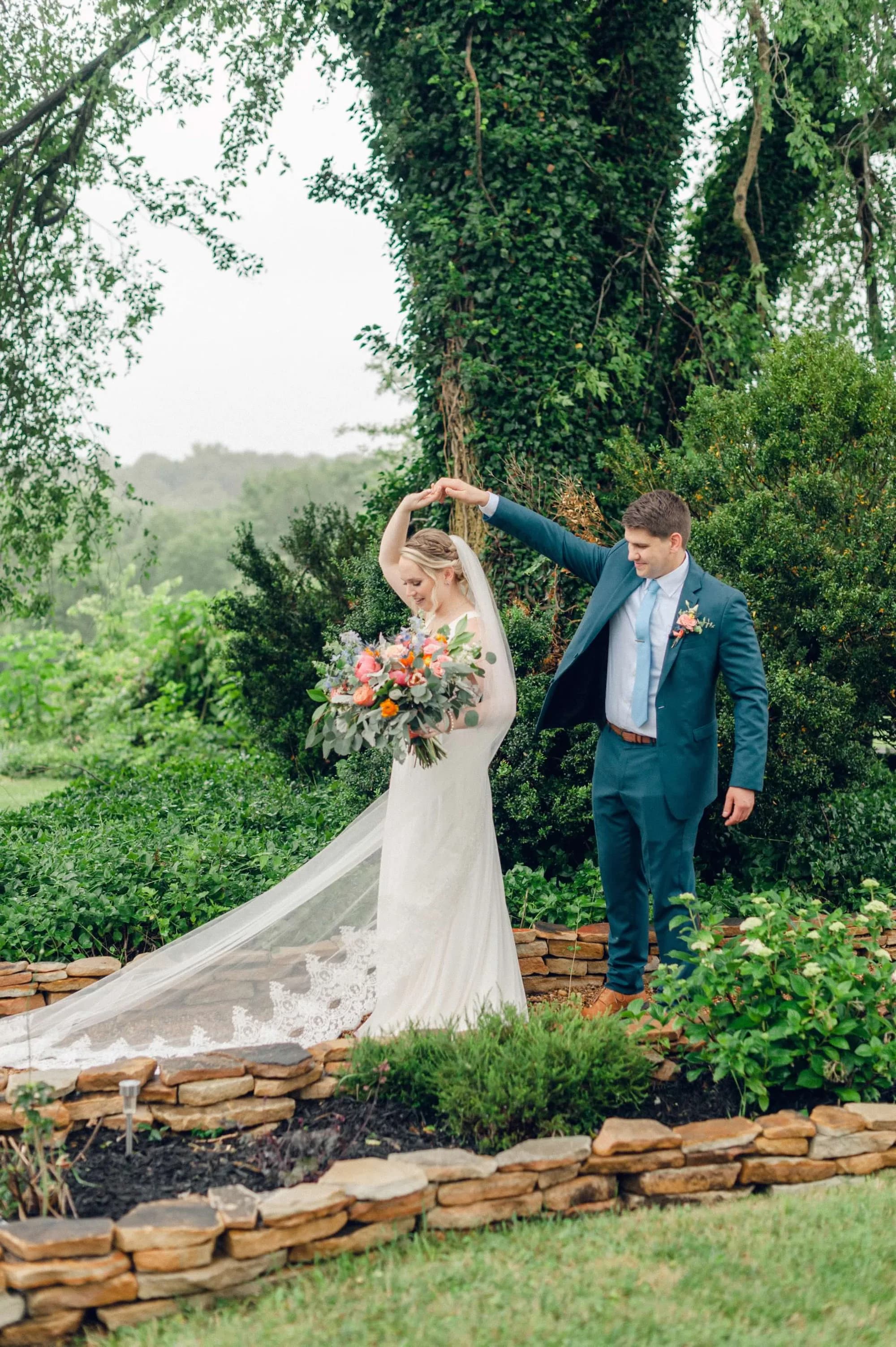 Groom twirls bride in lace gown among lush stone-bordered gardens at Rixey Manor, colorful bouquet in hand
