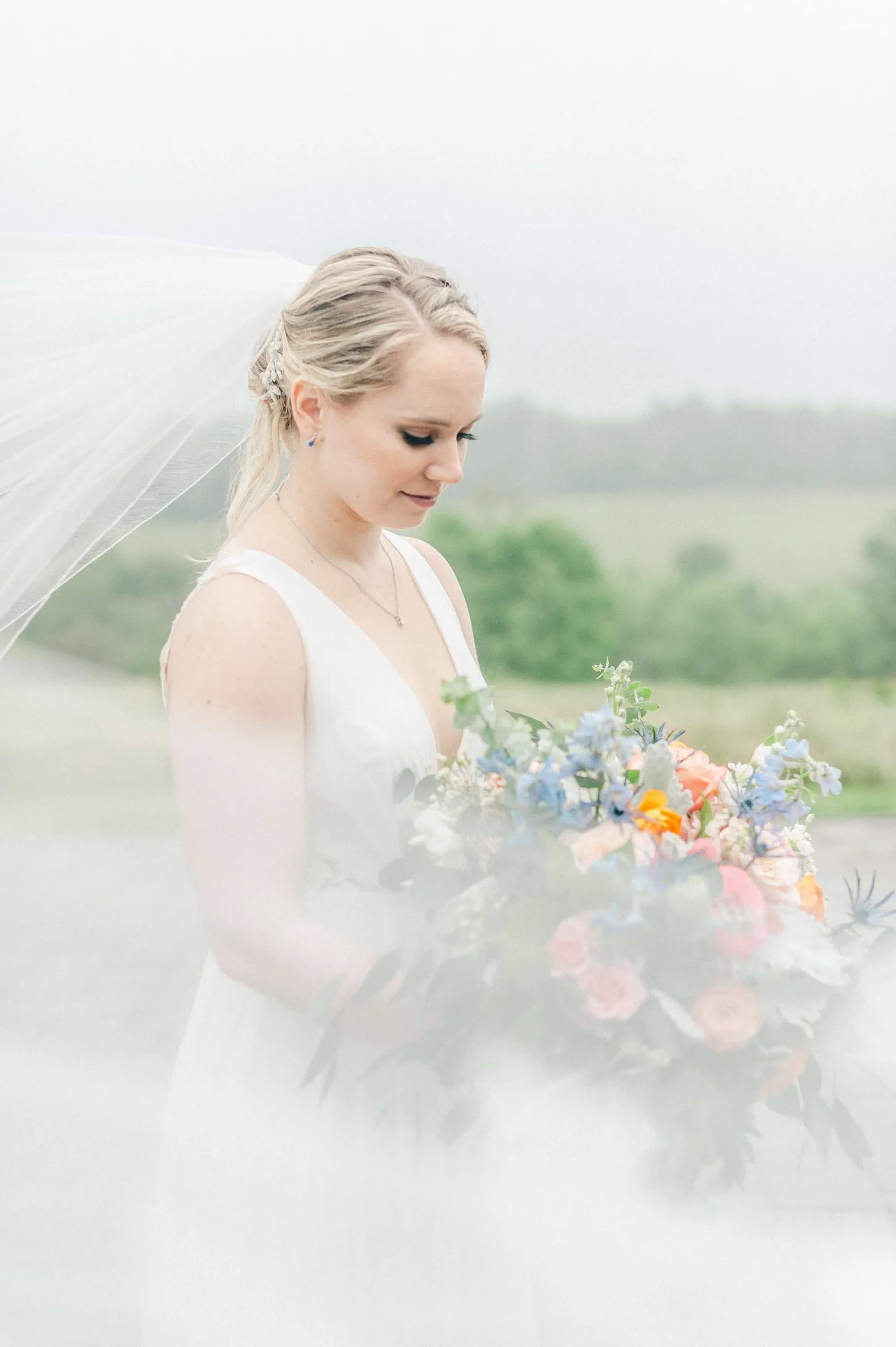 Bride in white gown holding colorful bouquet, veil billowing softly, gazing down in serene outdoor portrait