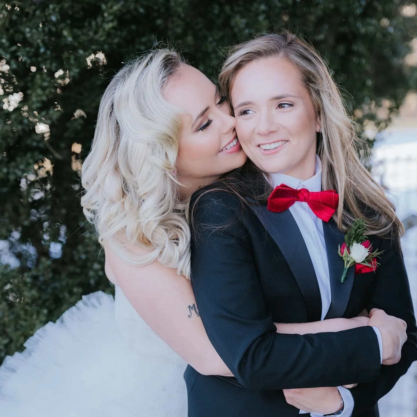 Two brides pose together outdoors, one in white dress kissing the other in black tuxedo with red bow tie.