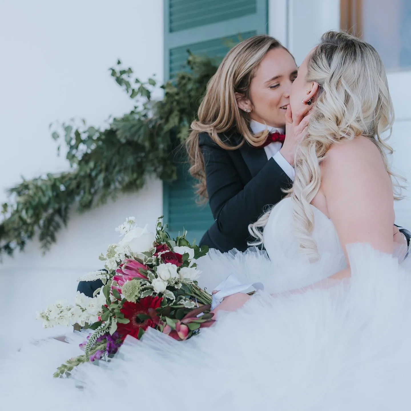 Two brides share a tender kiss, one in a tuxedo and one in a white gown, with a red and white bouquet nearby.
