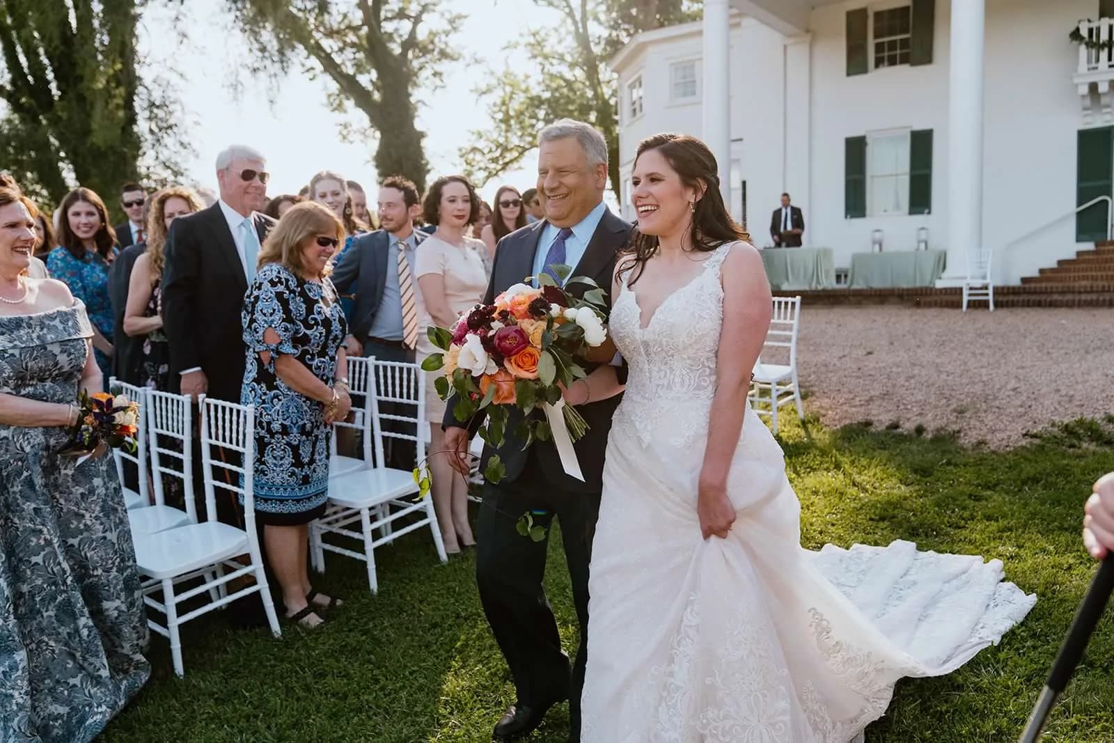 Bride walks down the aisle with her father at Rixey Manor, laughing, guests seated on white chairs on sunny lawn