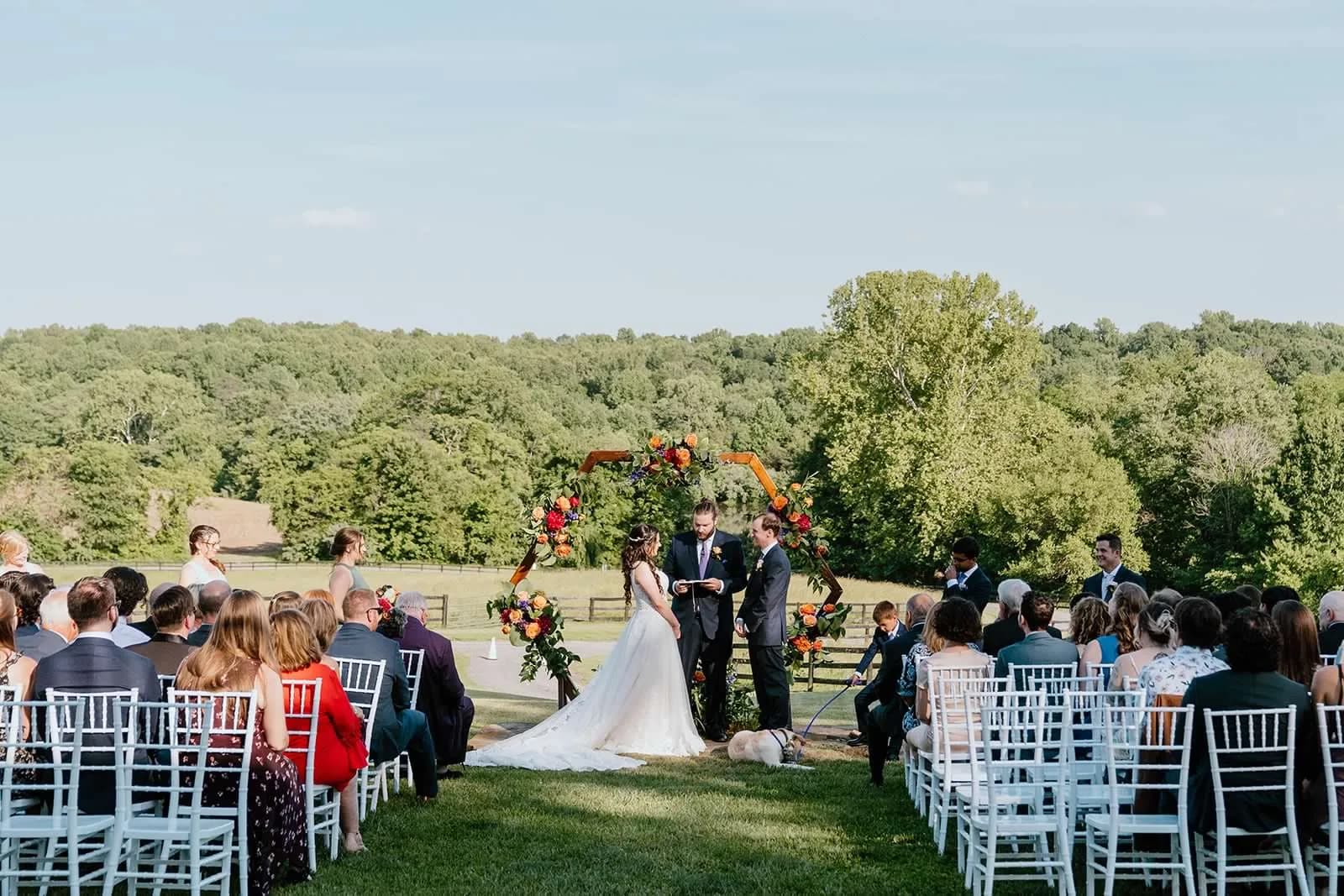 Outdoor wedding ceremony at rural venue with bride, groom, and guests seated in white chairs facing altar with floral arch.