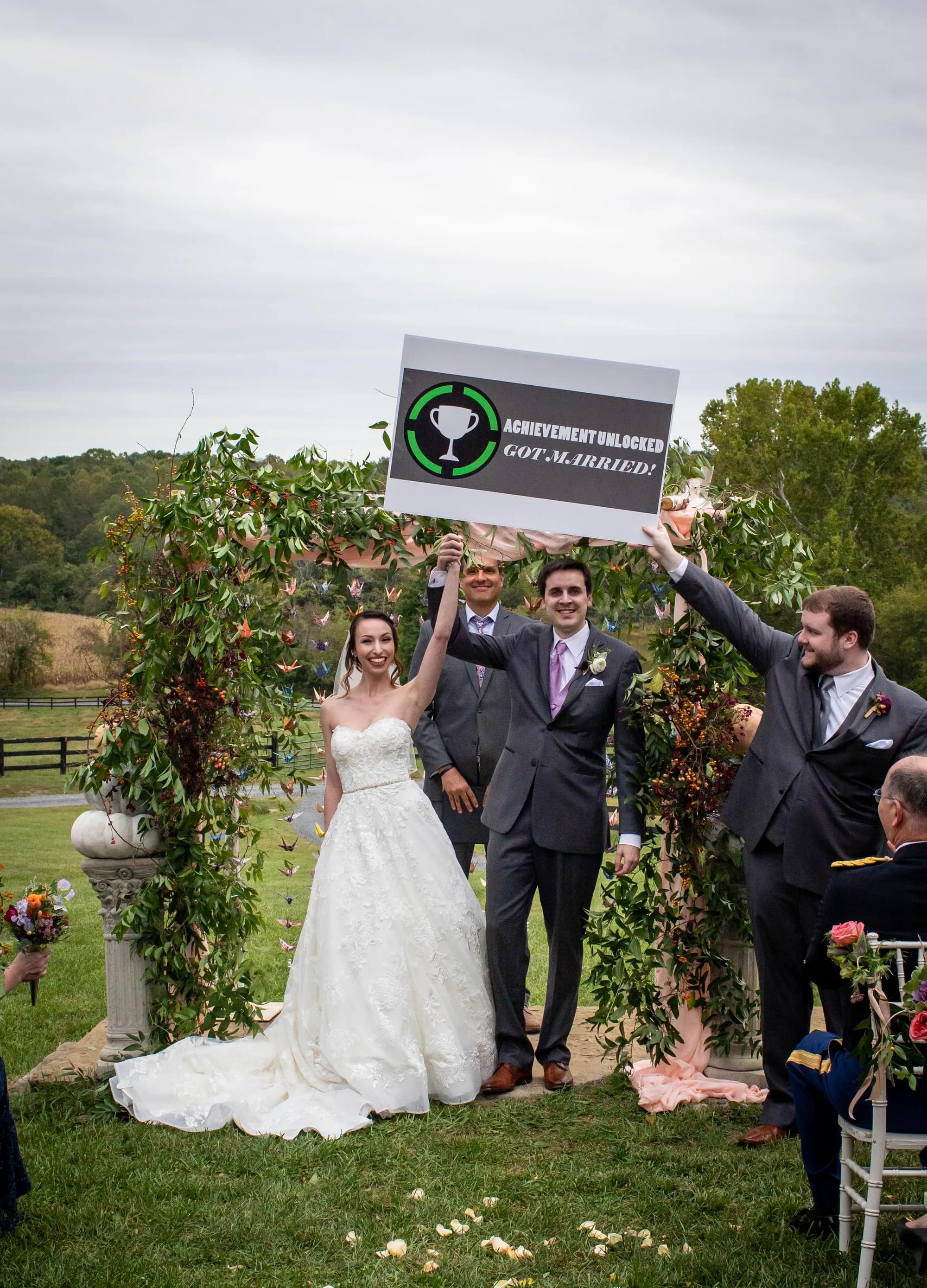 Newlyweds celebrate under floral arch at Rixey Manor holding 'Achievement Unlocked: Got Married' sign